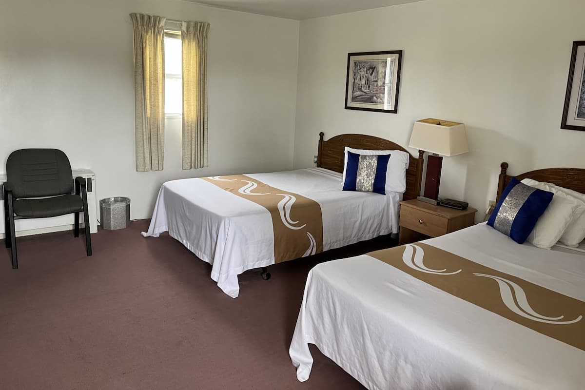 Ein Hotelzimmer mit zwei Betten, einem Sessel, einem Nachttisch, einer Lampe, einem Bild an der Wand und einem Fenster mit Vorhängen.