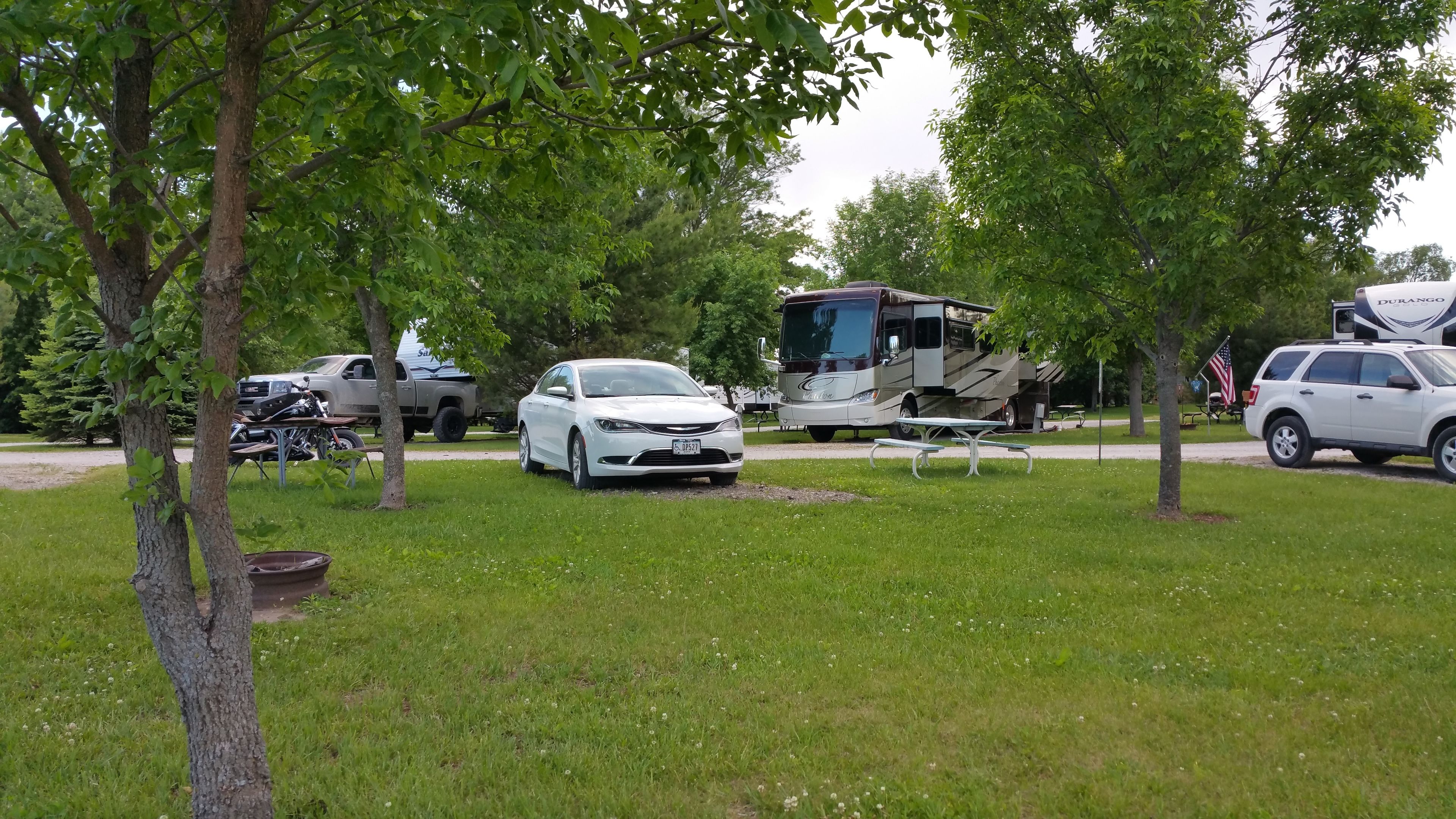 Un camping con vehículos recreativos, un coche blanco y mesas de picnic.