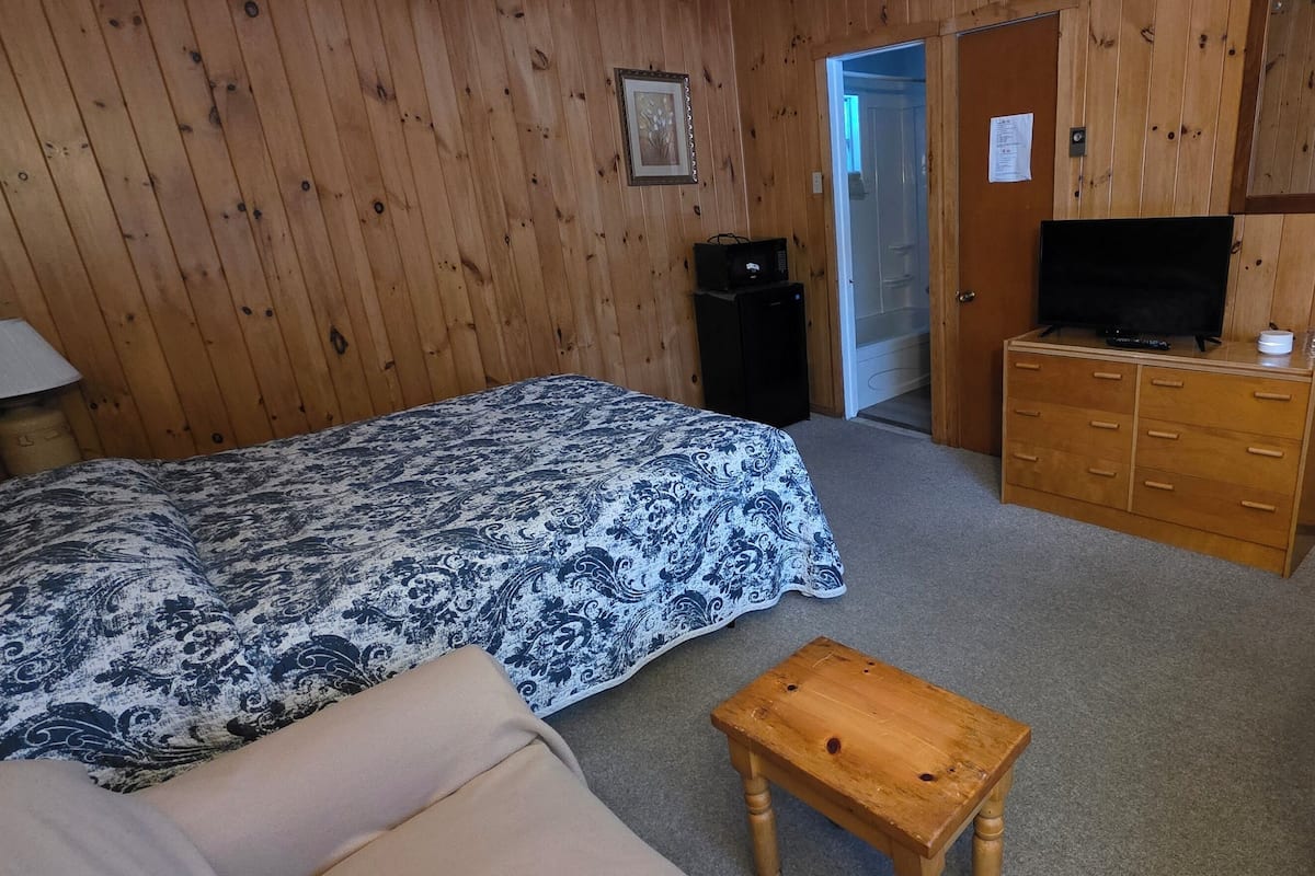 A room with wooden panel walls, a bed with a patterned bedspread, a wooden dresser with a television, and a small wooden table.