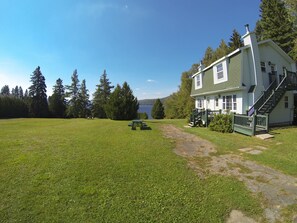 A house with a front porch, a staircase, and a picnic table in a grassy area with trees and a lake in the background. - Motel Le Fleuron (Saint Donat)