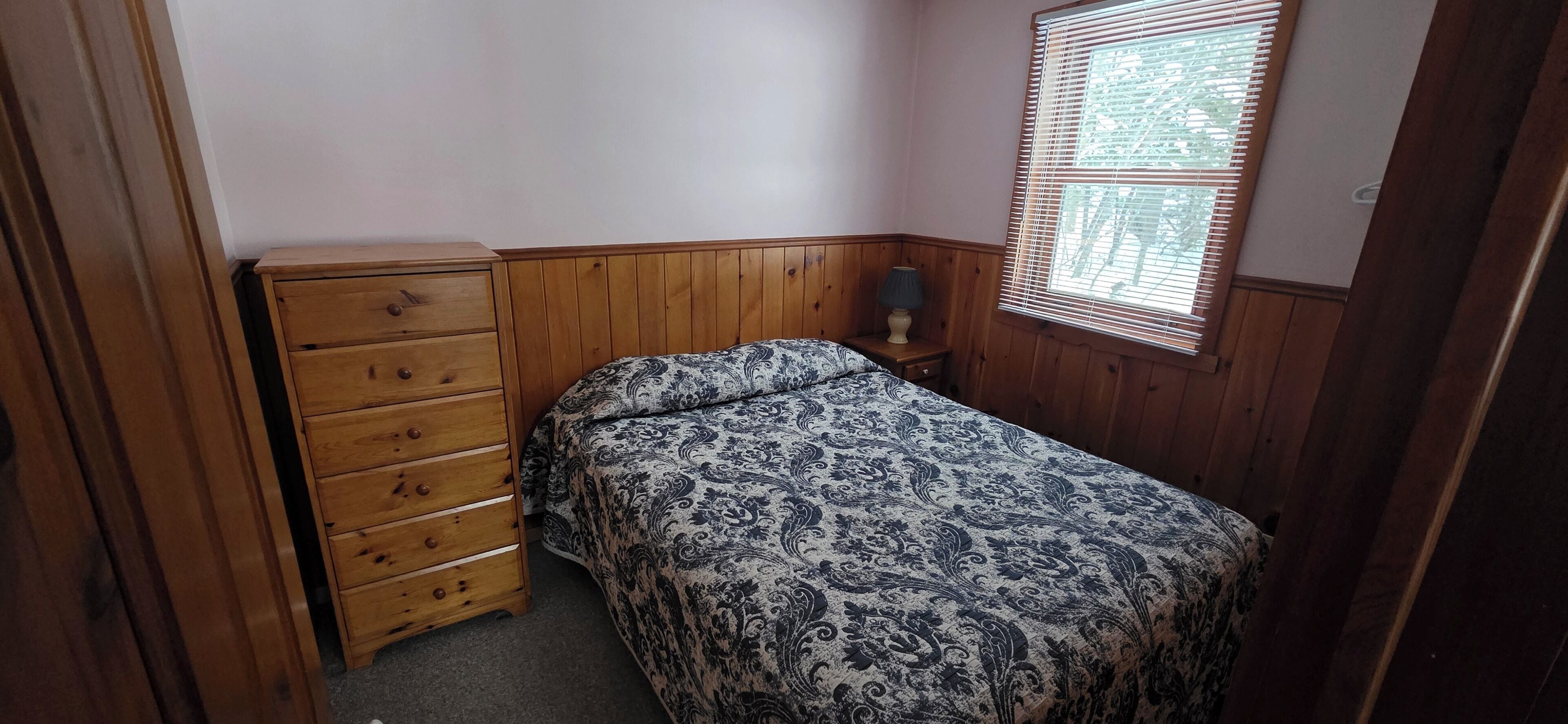 Ein Schlafzimmer mit einem Holzkapuzen, einem Bett mit gemustertem Bettbezug, einem Nachttisch mit Lampe und einem Fenster mit Jalousien.