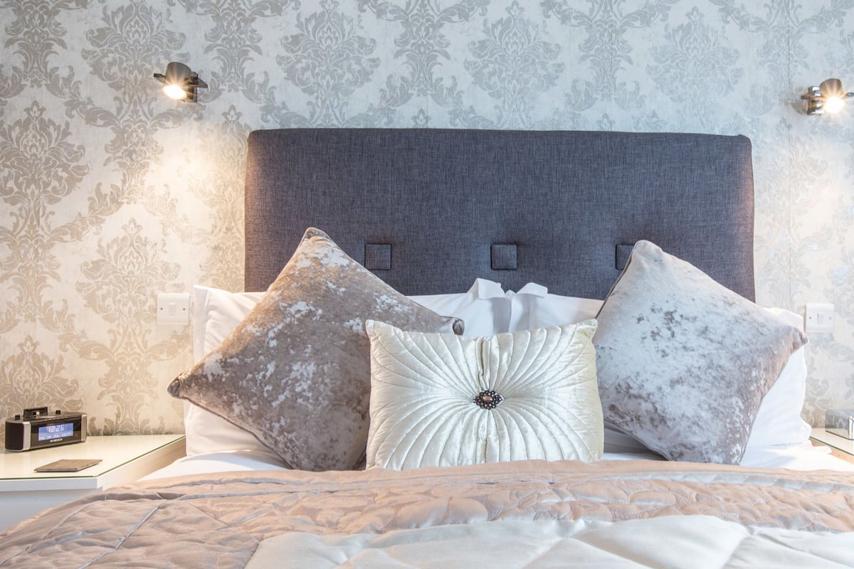 A neatly made bed with a grey headboard, two bedside tables with lamps, and a patterned wallpaper.