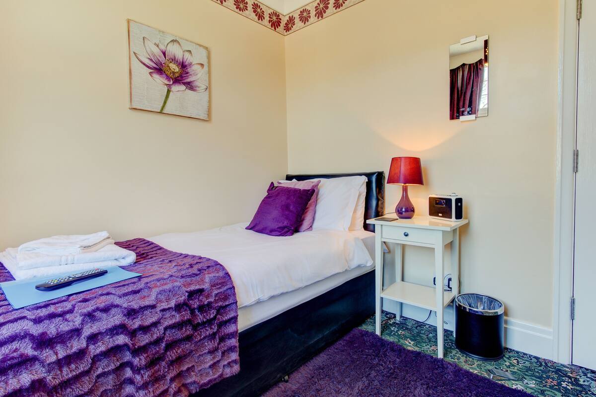 A neatly made bed with a purple blanket and pillows, a bedside table with a lamp and a small electronic device, a trash can, and a wall-mounted mirror.