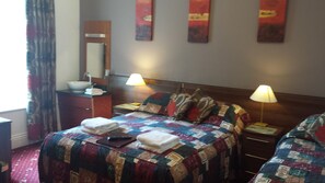 A hotel room with two beds, a bedside table with a lamp, a nightstand, a bathroom with a sink and mirror, and a window with curtains. - Karden House (Llandudno)