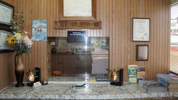 Un comptoir d’accueil avec un mur recouvert de panneaux en bois, un certificat encadré, un panneau indiquant « Merci de ne pas fumer », un vase avec des fleurs et une enseigne « Lincoln Motel ».