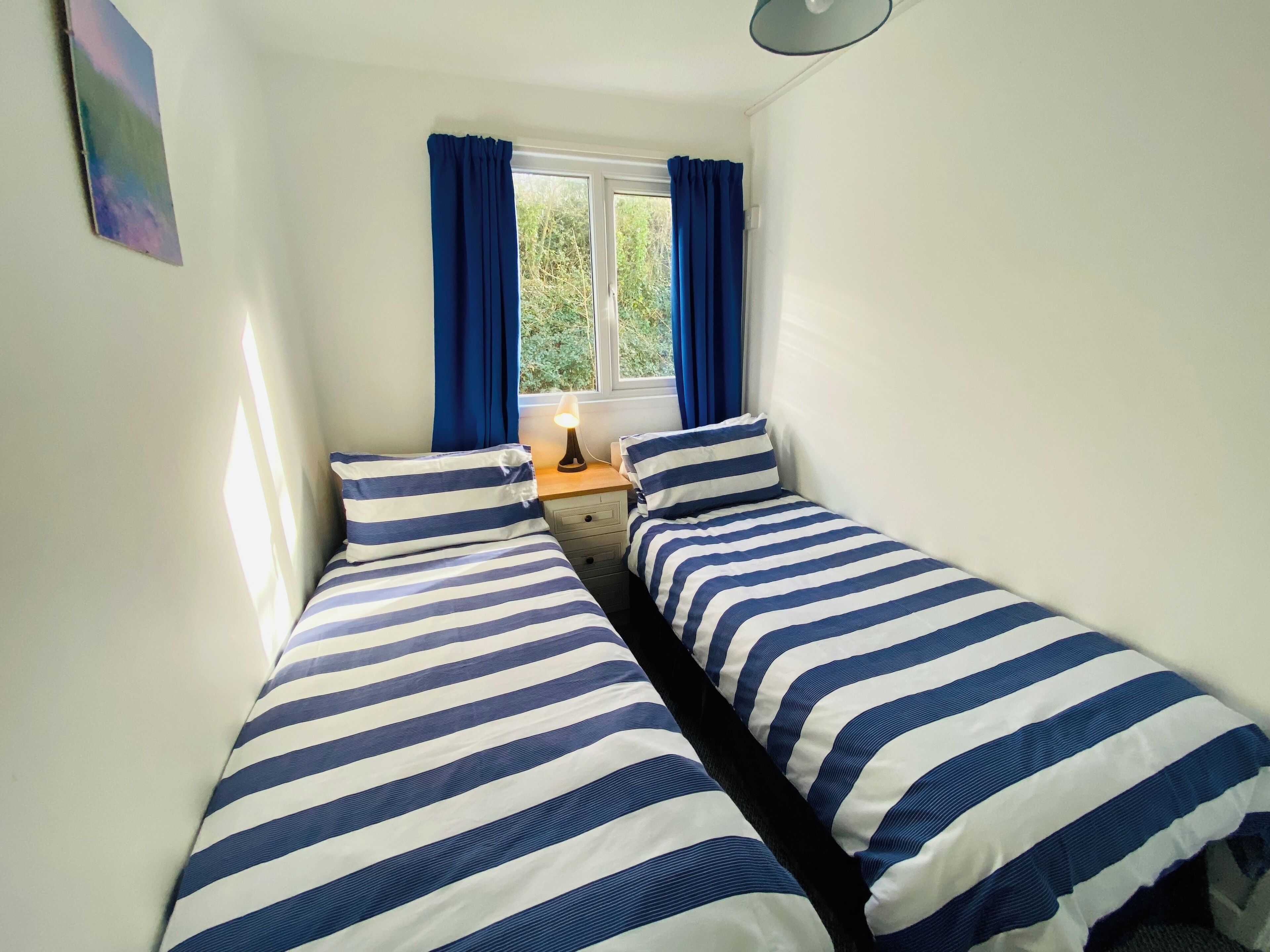 A small, well-lit room with two single beds, a nightstand, a lamp, and a window with curtains.