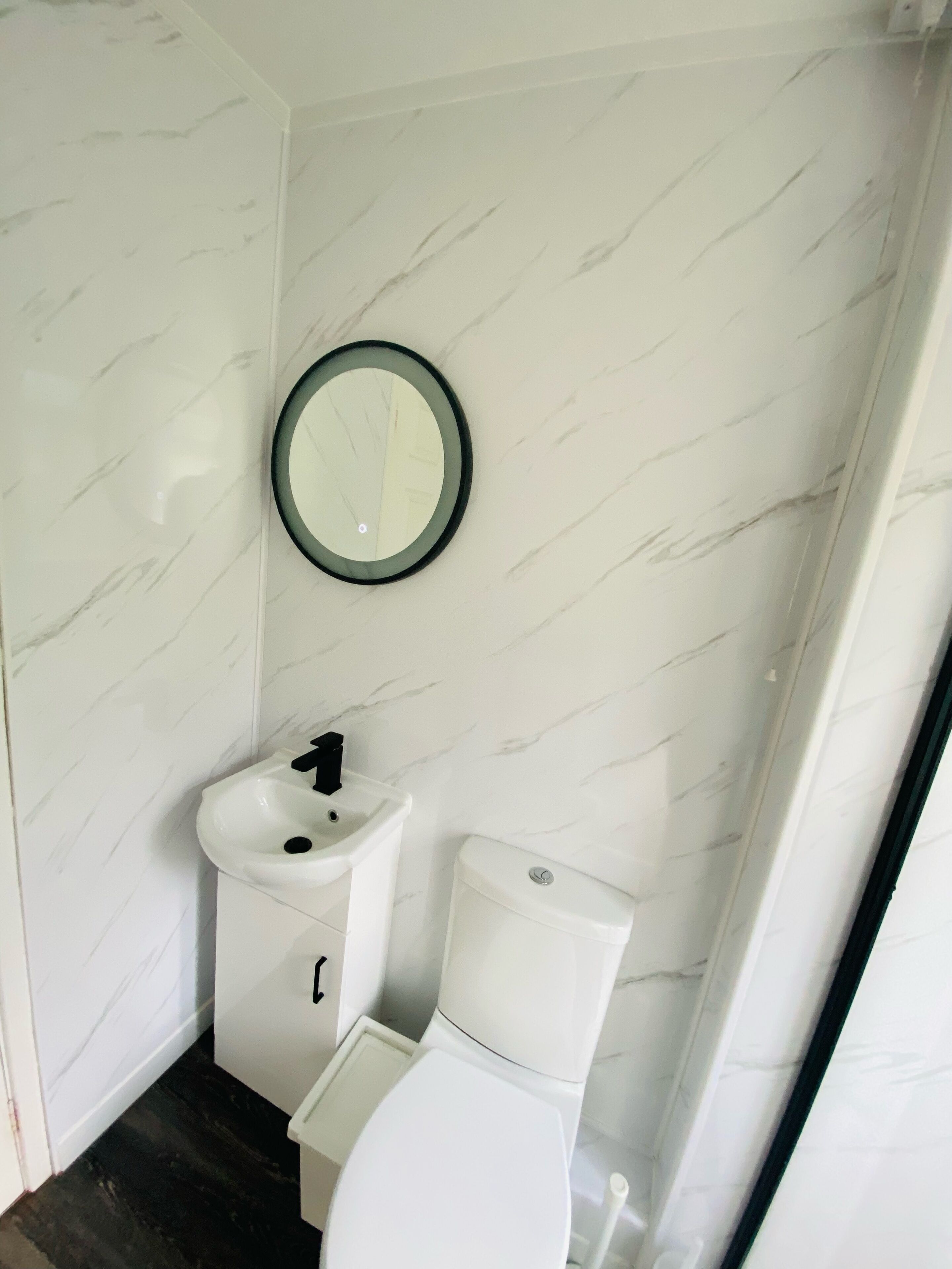 Et badeværelse med en hvid marmorvæg, et rundt spejl, en hvid vask og et toilet.