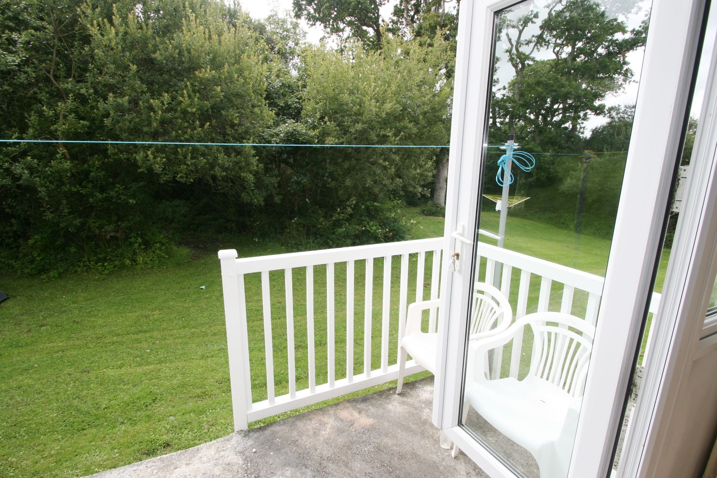 Un balcón con barandilla blanca, una silla de plástico blanca y vista a un jardín con árboles.