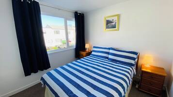 Un dormitorio con cama, mesita de noche, lámpara, ventana con cortinas e imagen enmarcada en la pared.