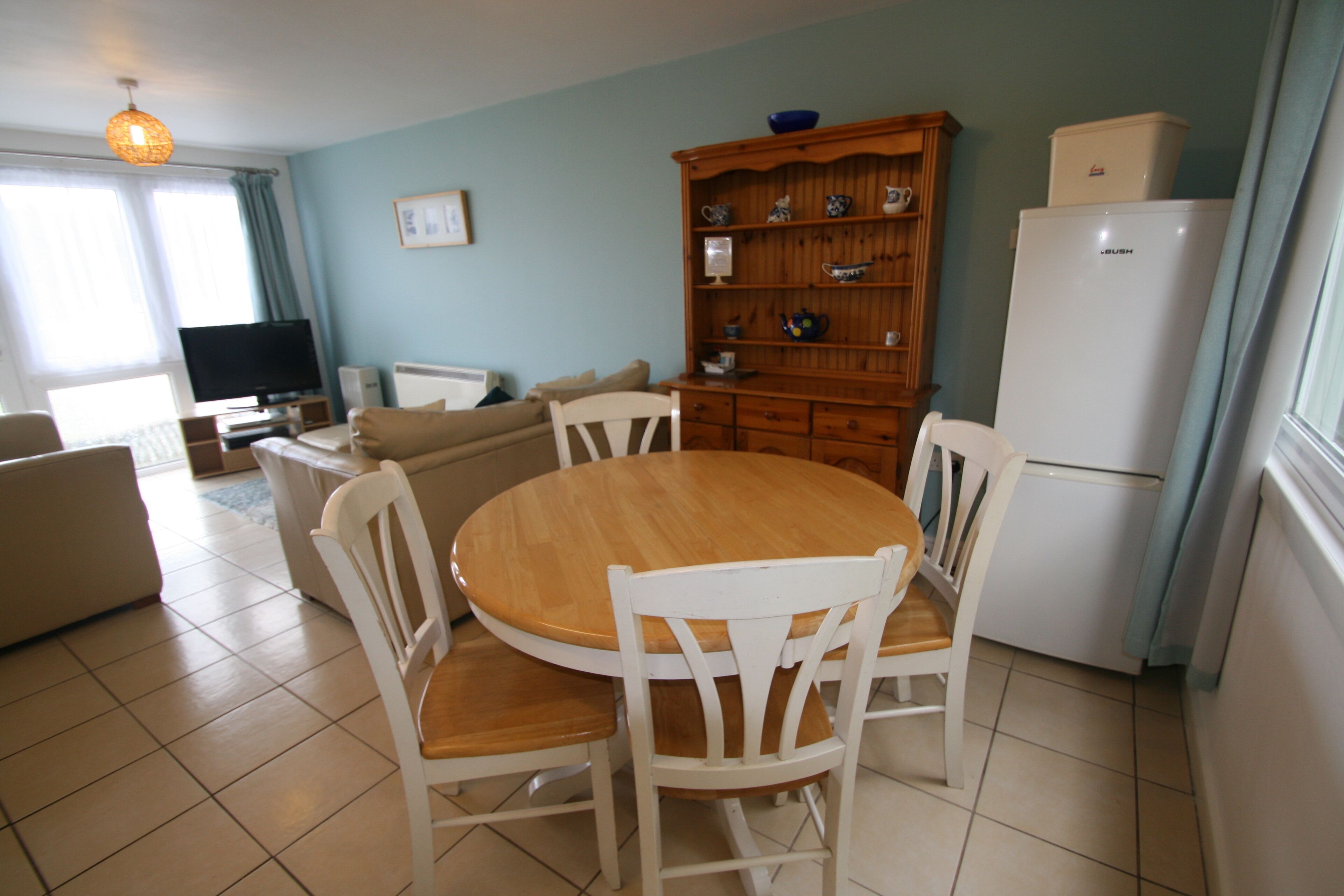 Un coin repas avec une table ronde en bois et des chaises blanches, un buffet en bois contenant de la vaisselle, un téléviseur et un réfrigérateur.