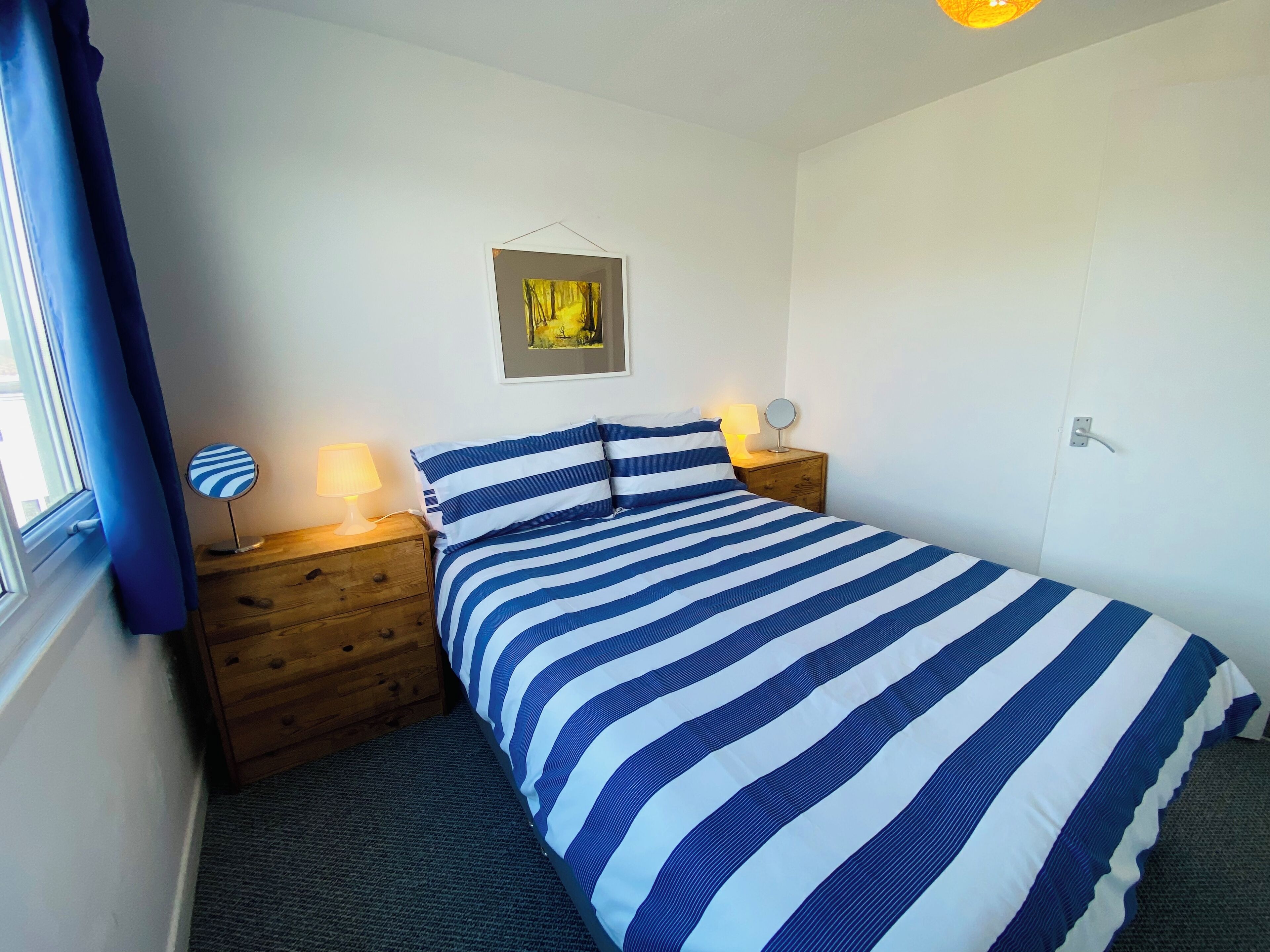 Quarto com cama, mesinhas de cabeceira de madeira, abajur, quadro na parede e janela com cortinas.