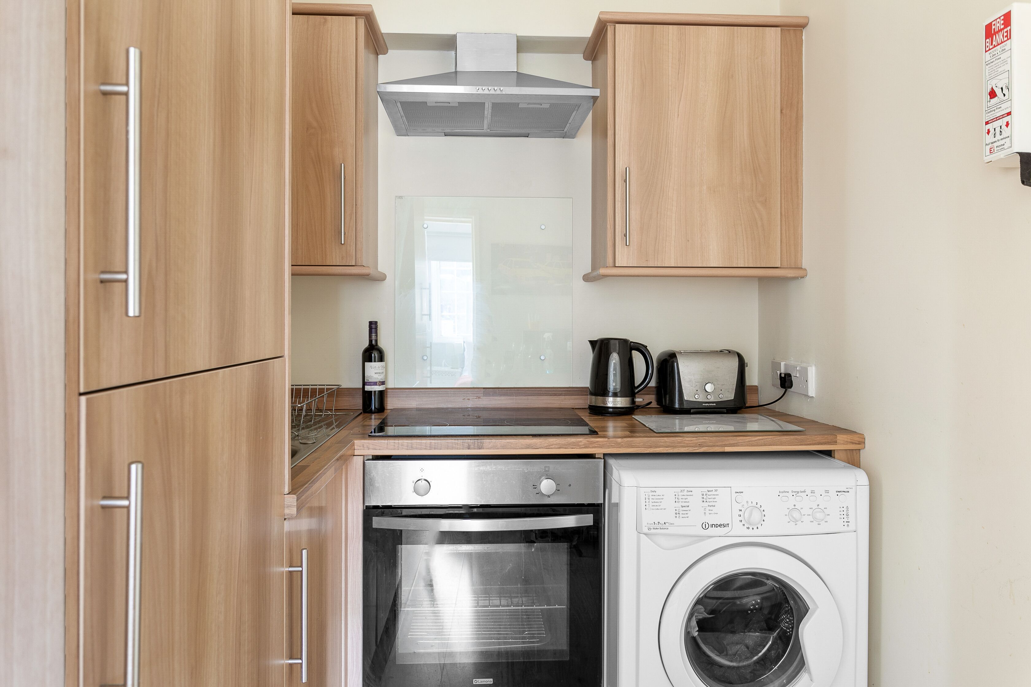 A compact kitchen with wooden cabinets, a built-in oven, a washing machine, and a toaster.