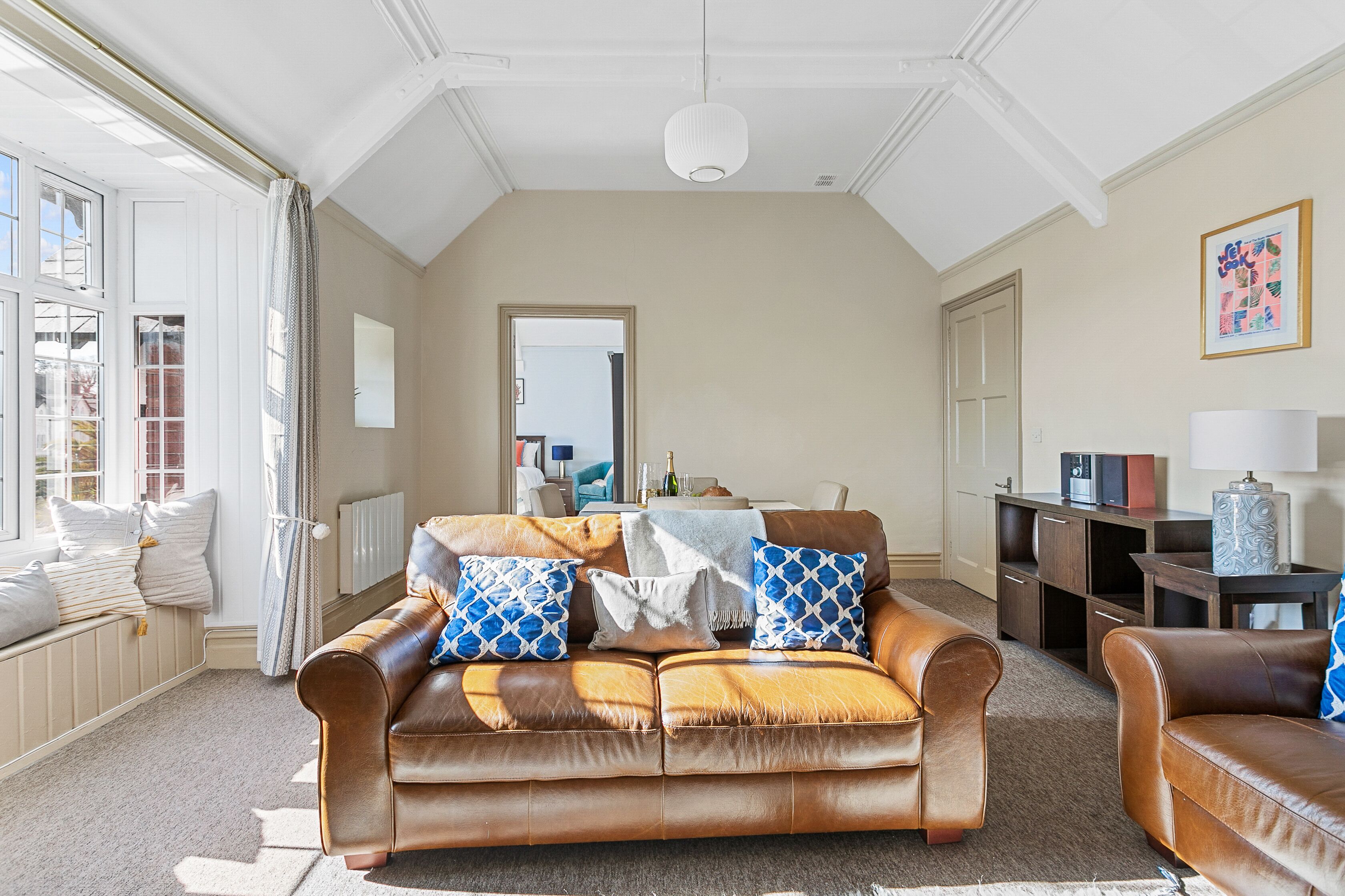 A spacious living room with a brown leather sofa, a coffee table, and a view of the outside through large windows.