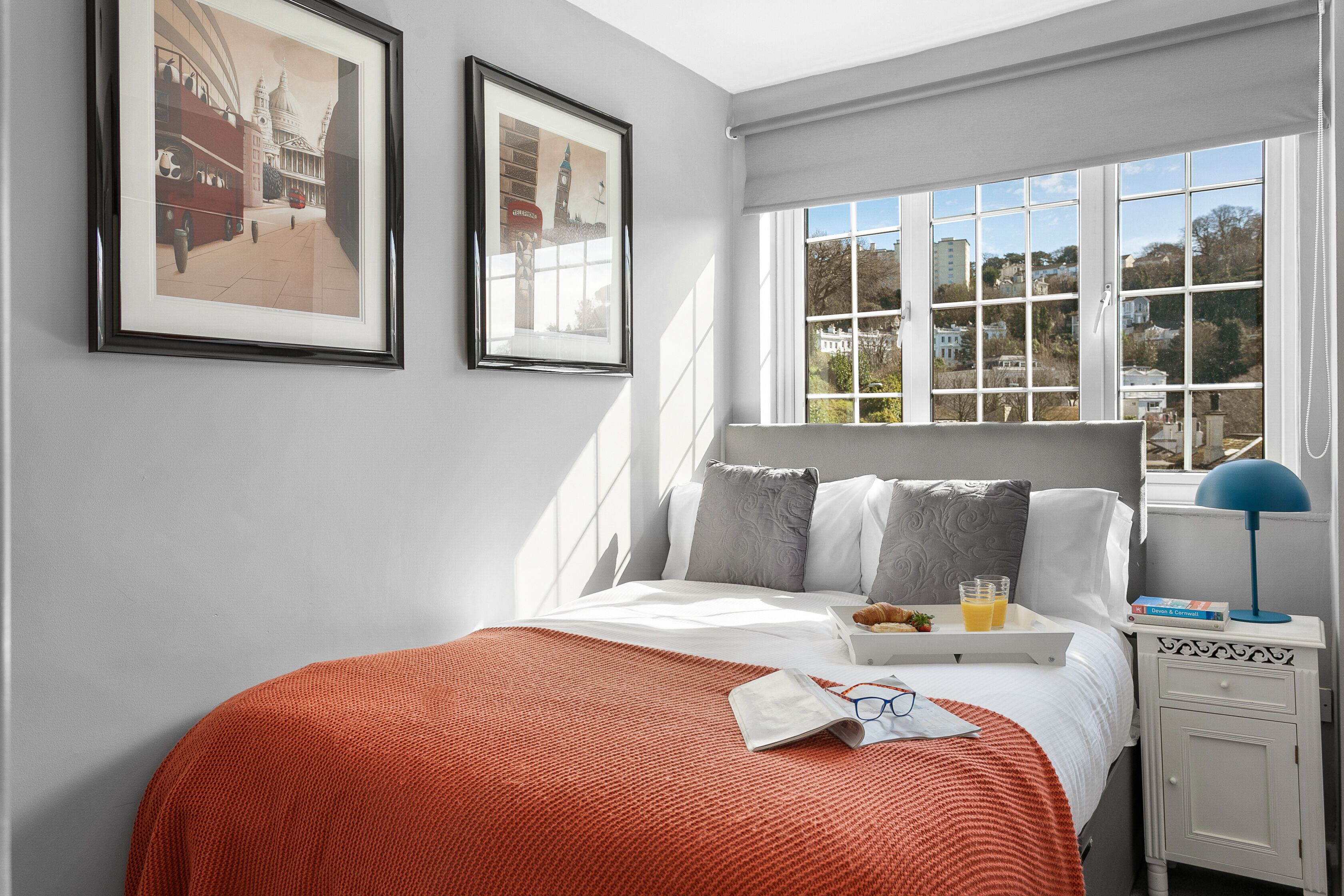 A neatly arranged bedroom with a large bed, a bedside table with a lamp, and a window with a view of buildings.