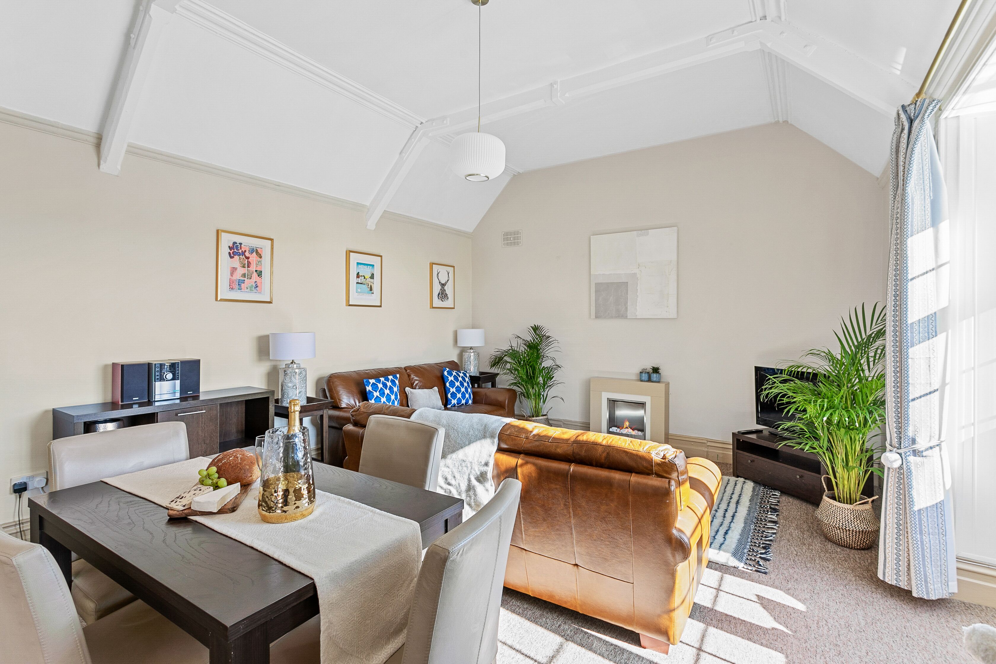 A spacious living room with a brown sofa, a dining table with a bottle and grapes, and a TV in the background.