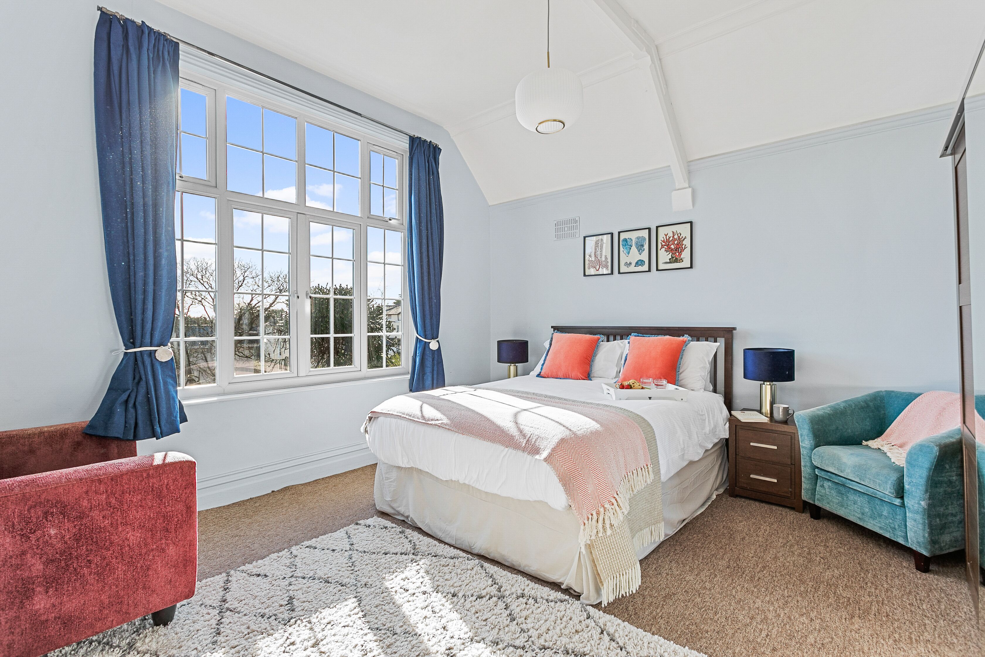 A bedroom with a large window, a bed with multiple pillows, a nightstand, a chair, and a rug.