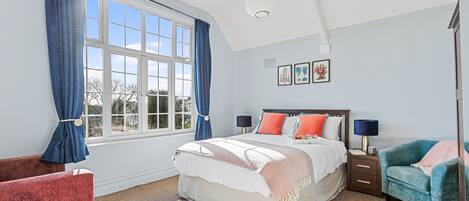 A bedroom with a large window, a bed with multiple pillows, a nightstand, a chair, and a rug.
