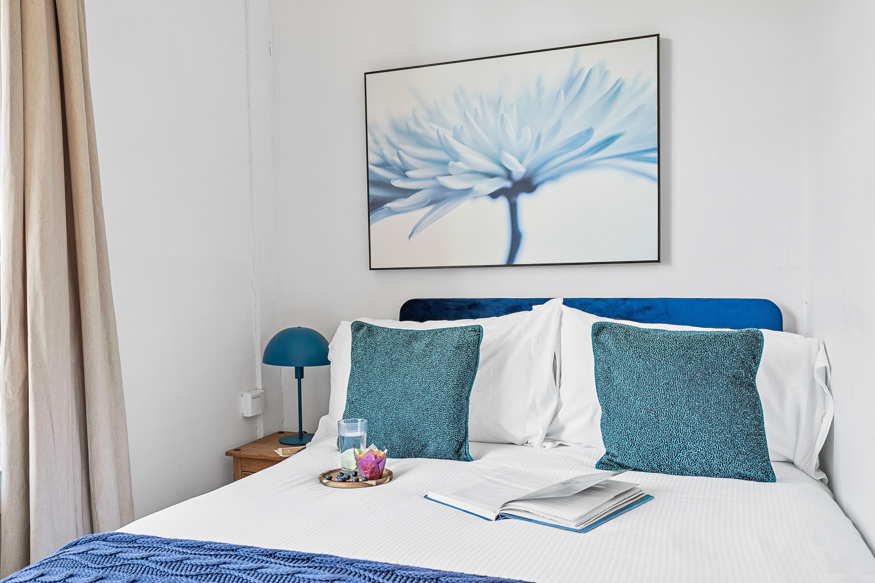 A neatly made bed with a blue and white pillow, a framed artwork of a flower, and a bedside table with a lamp and a glass of water.