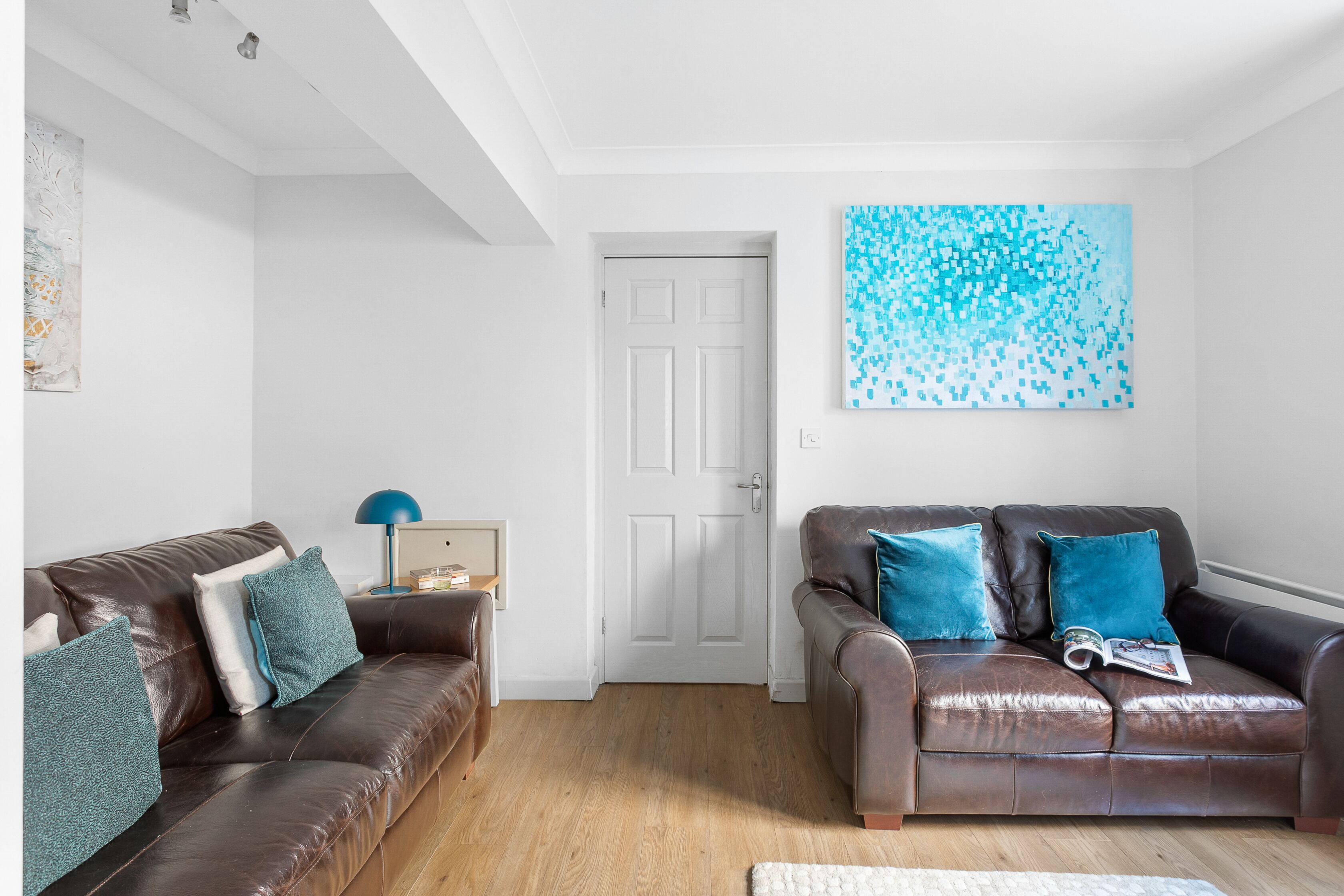 Een woonkamer met twee bruine leren banken, een witte deur en een blauw abstract schilderij aan de muur.