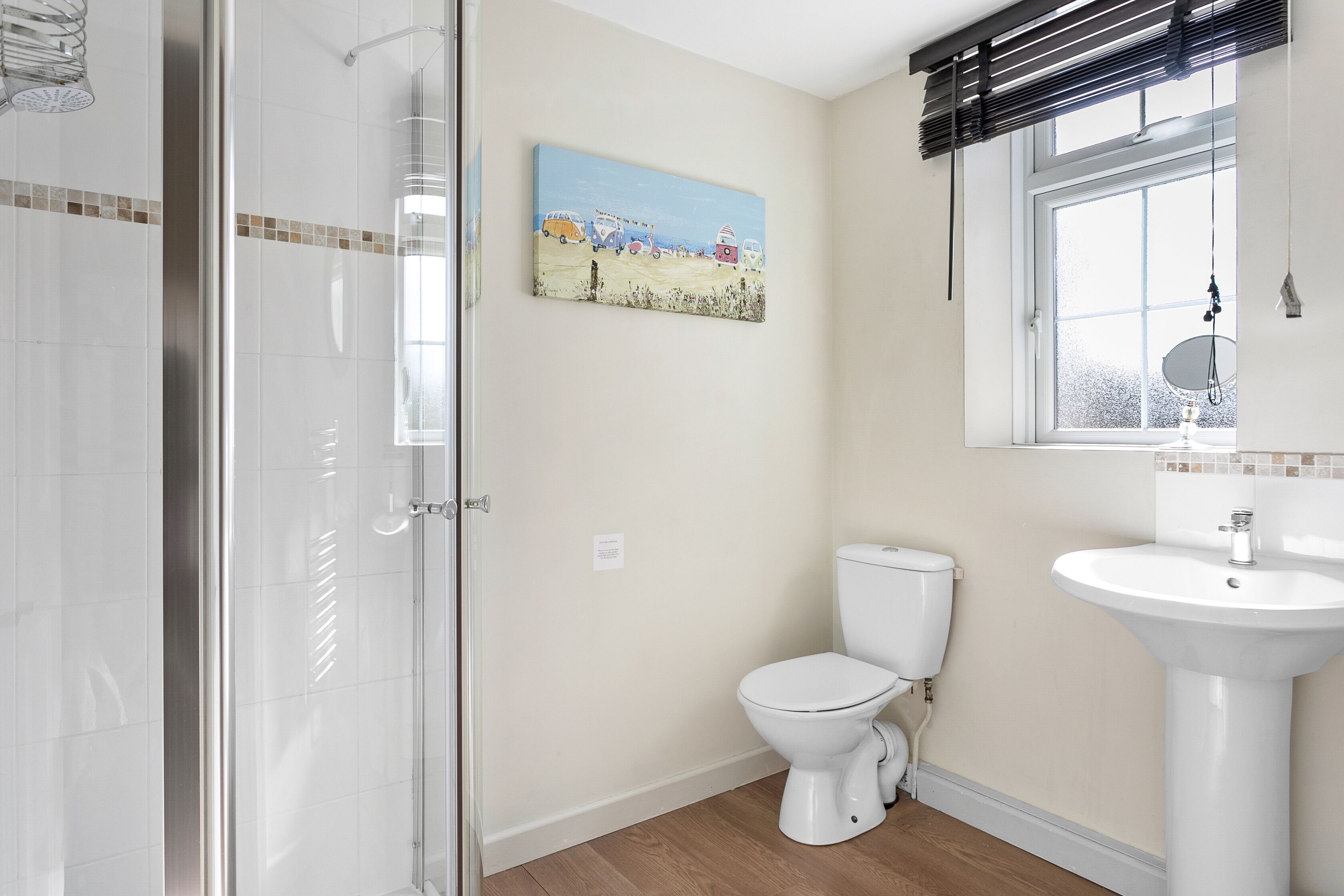 A bathroom with a glass shower door, a toilet, a sink, and a window with blinds.