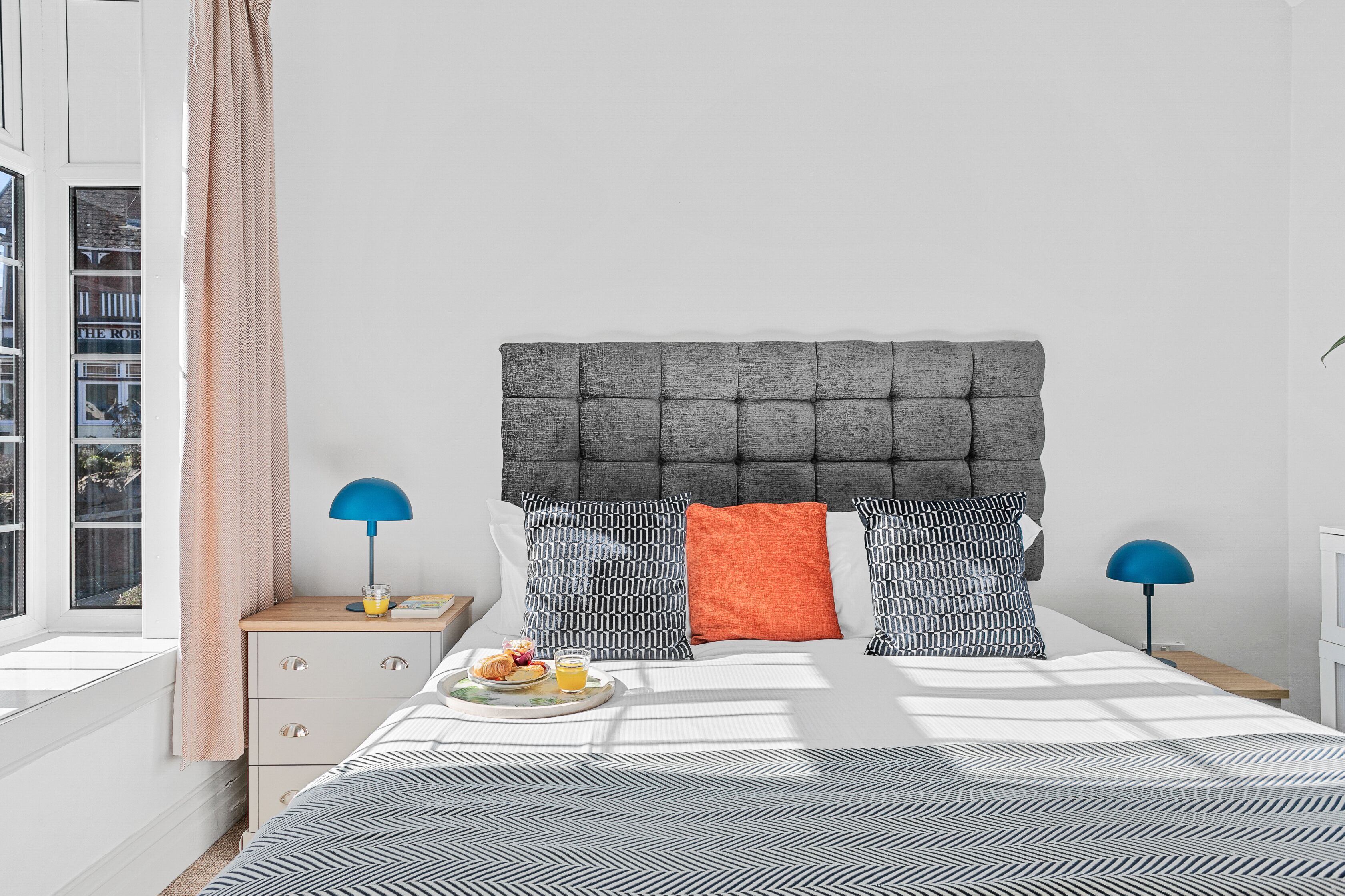 Une chambre à coucher avec une tête de lit capitonnée, une literie à motifs, une table de chevet avec une lampe et un plateau de petit-déjeuner sur le lit.