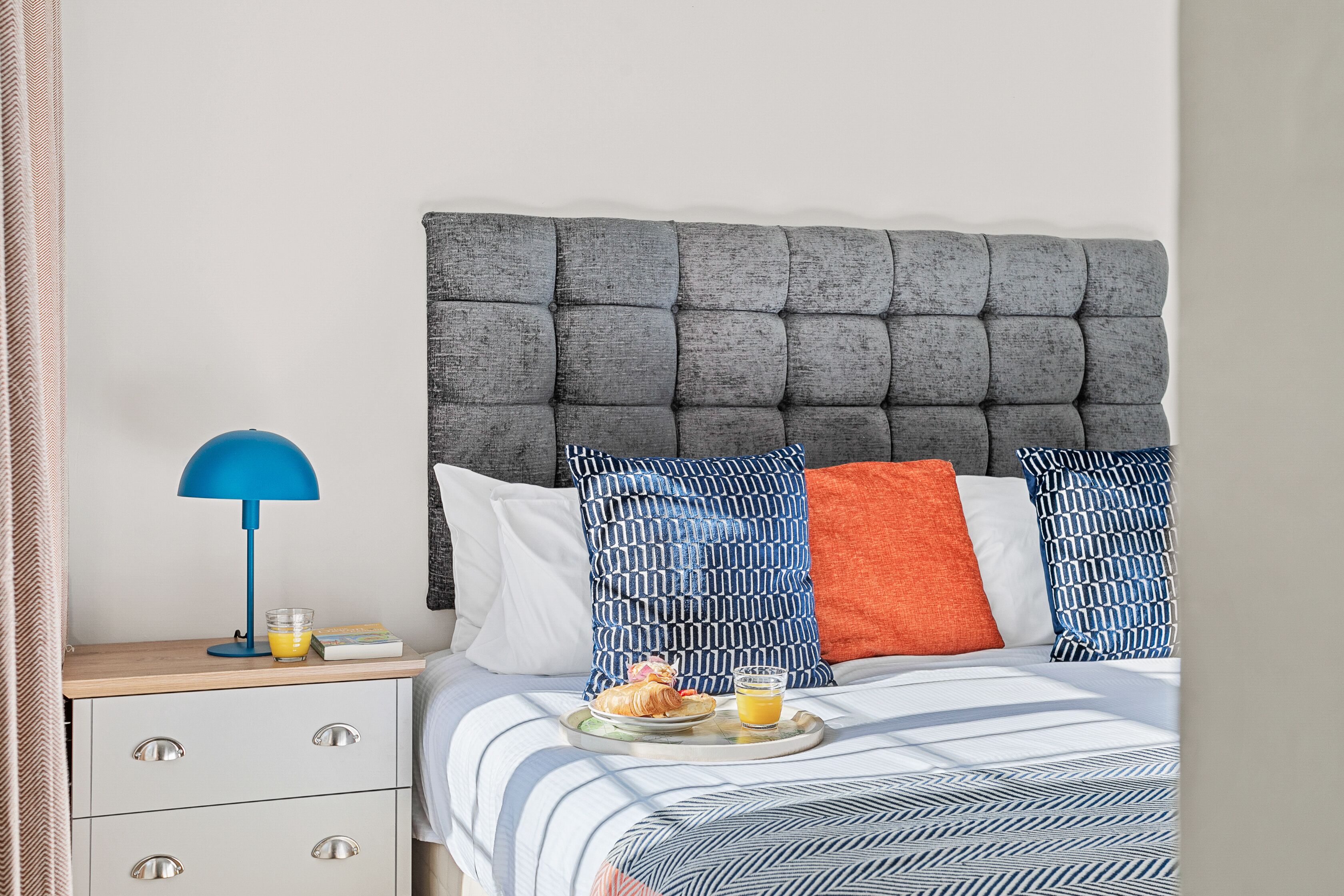 A neatly made bed with a grey tufted headboard, a blue and white patterned pillow, and an orange pillow. A bedside table holds a blue lamp, a glass of orange juice, and a tray with croissants and a small bowl of jam.