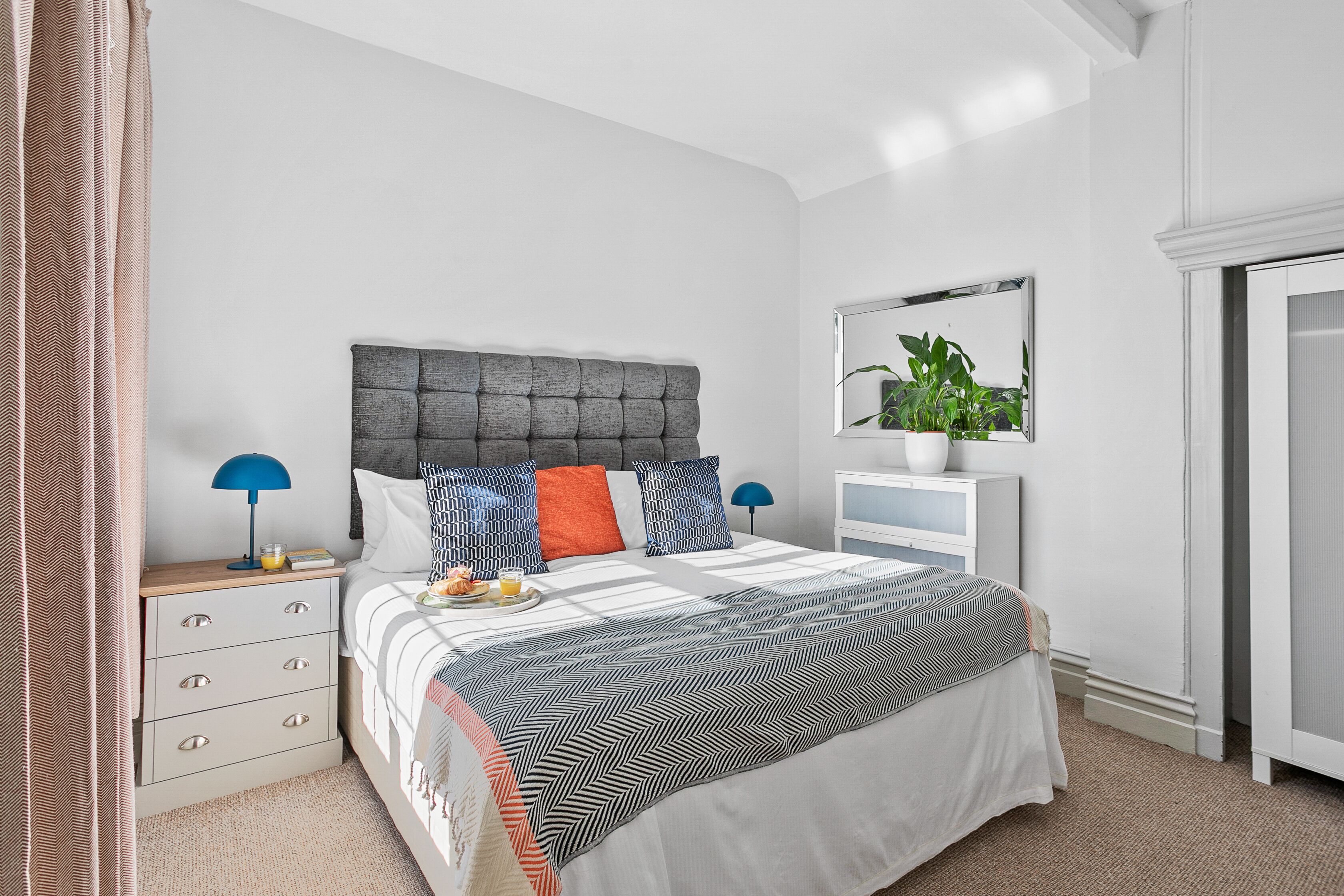 A neatly arranged bedroom with a large bed, a bedside table with a lamp, a dresser, and a mirror on the wall.
