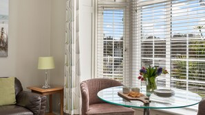 Ett modernt vardagsrum med ett glasbord, en brun lädersoffa, ett litet sidobord med en lampa och ett fönster med persienner.
