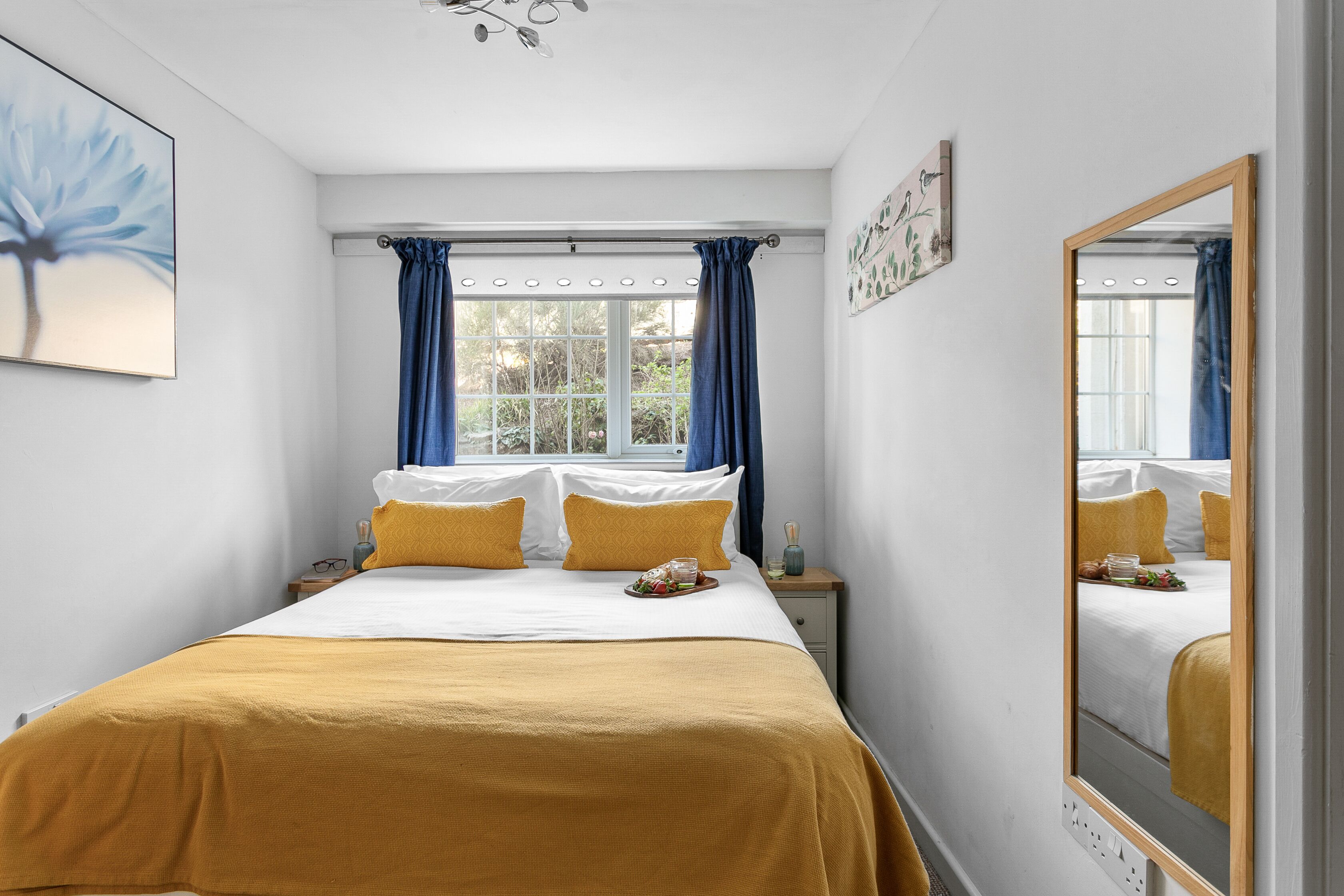 Cama bem arrumada com colcha amarela, duas almofadas e uma bandeja de frutas. Há uma janela com cortinas azuis, uma mesa de cabeceira com abajur e um espelho na parede.