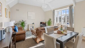 Ett rymligt vardagsrum med en öppen spis, lädersoffor, ett matbord med stolar och ett stort fönster med gardiner.