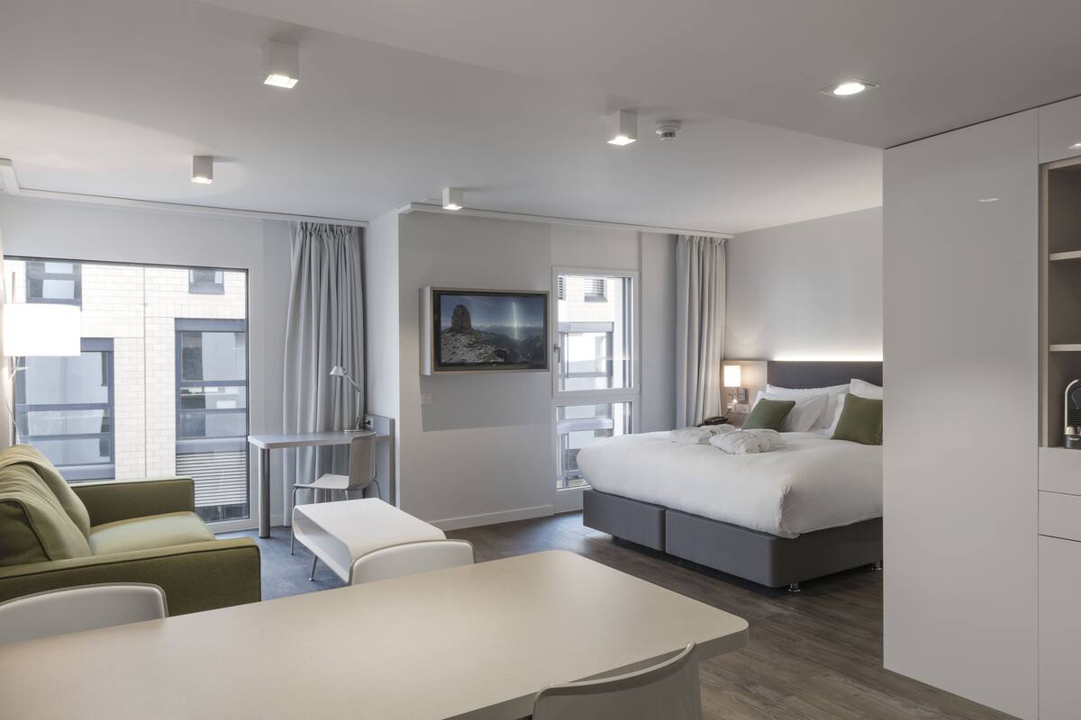 A modern hotel room with a bed, a desk, a chair, and a view of the cityscape.