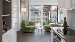 A modern kitchen with white cabinets, a dining area with a green sofa, and a view of the city through large windows. - Base Nyon (Nyon)