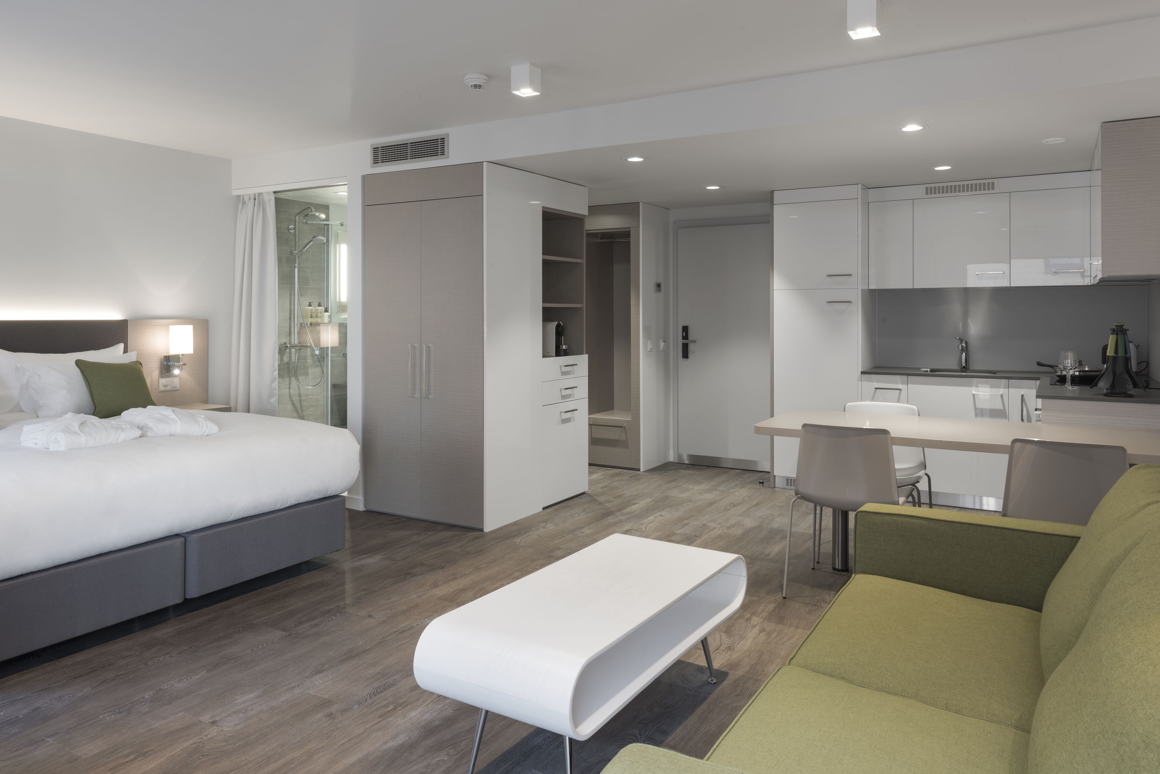 A modern hotel room with a bed, a bench, a sofa, a kitchenette, and a walk-in closet.