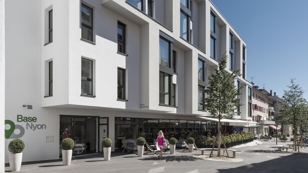 A modern hotel with a white facade, large windows, and a covered outdoor seating area with potted plants. - Base Nyon (Nyon)