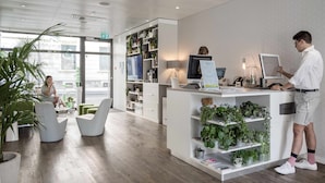 A modern reception area with a desk, computer, and shelves with plants. There are seating areas with chairs and a large window. - Base Nyon (Nyon)