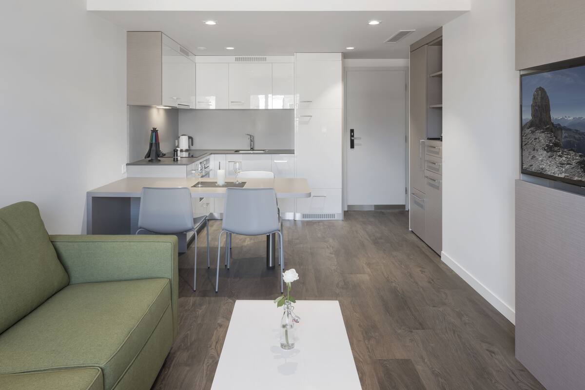A modern kitchen with white cabinets, a dining area with a green sofa and two chairs, and a vase with a single white rose on a white table.