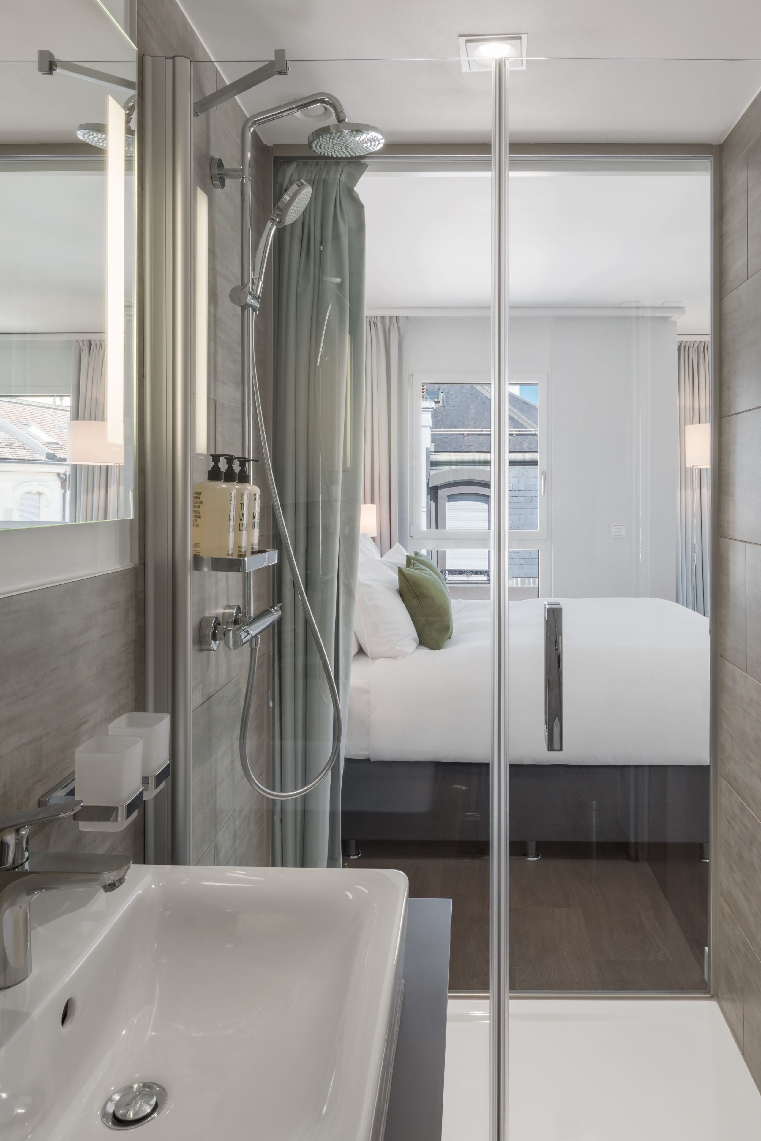 Ein modernes Hotelzimmer mit Duschkabine aus Glas, einem großen Bett mit weißer Bettwäsche, einem Nachttisch und Blick auf die Stadt durch das Fenster.