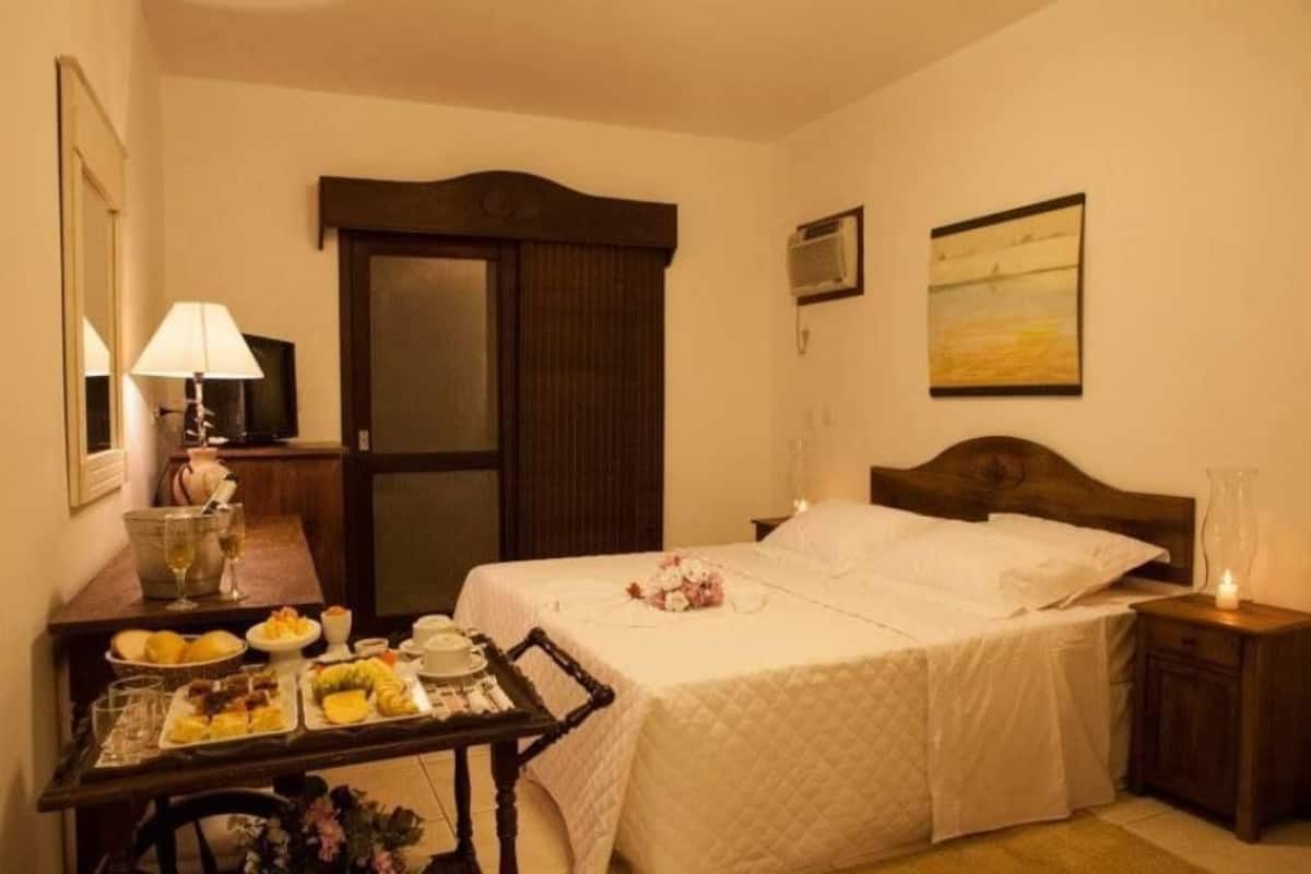Habitación de hotel con una cama, una mesita de noche con lámpara, una pequeña mesa con artículos de desayuno, un cuadro en la pared y un equipo de aire acondicionado.