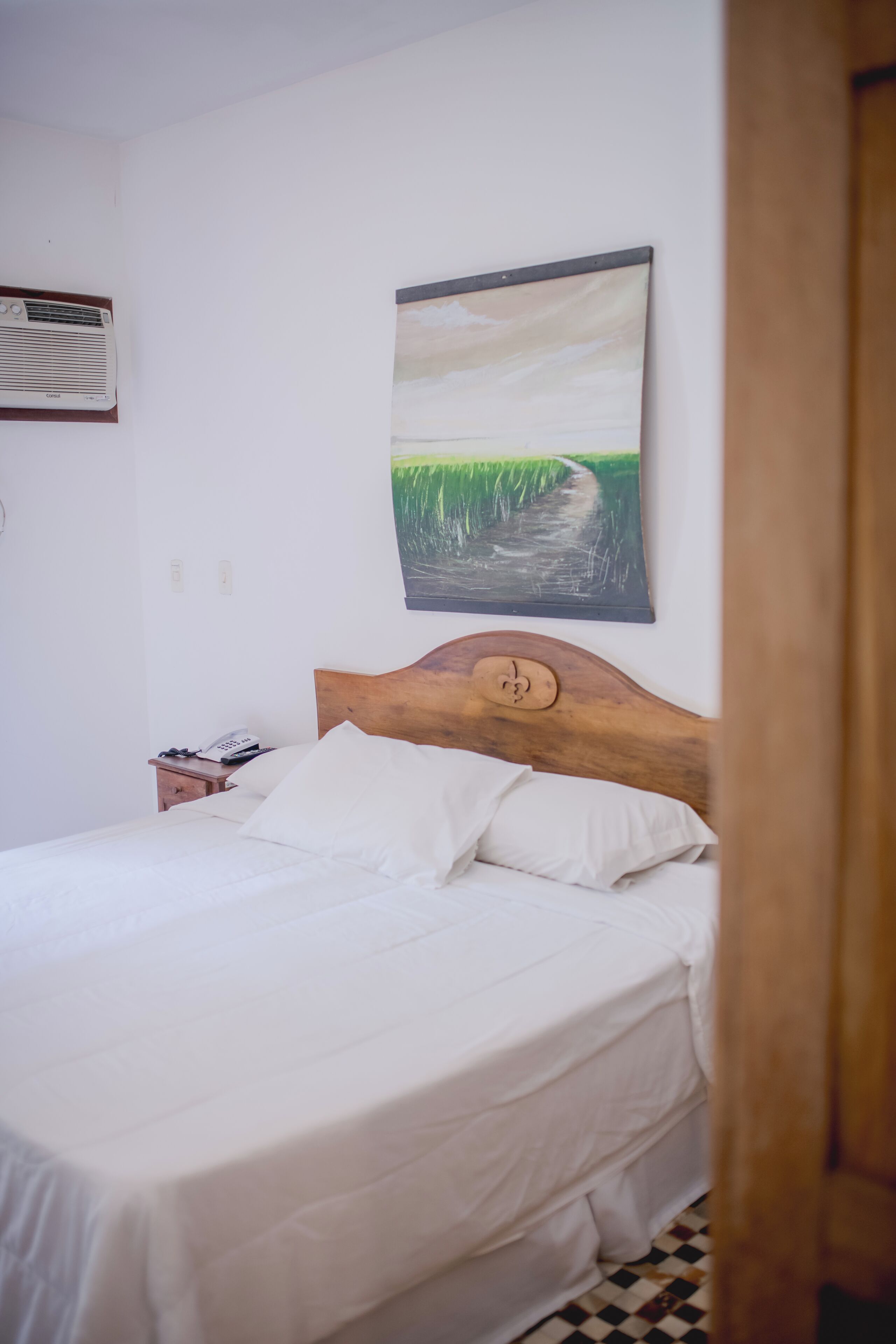 Una cama bien hecha con sábanas blancas, un cabecero de madera y un cuadro de un paisaje sobre ella.