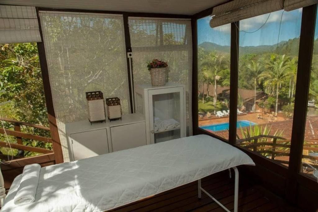 Quarto com cama, um armário pequeno branco e janela com vista para uma piscina e vegetação.