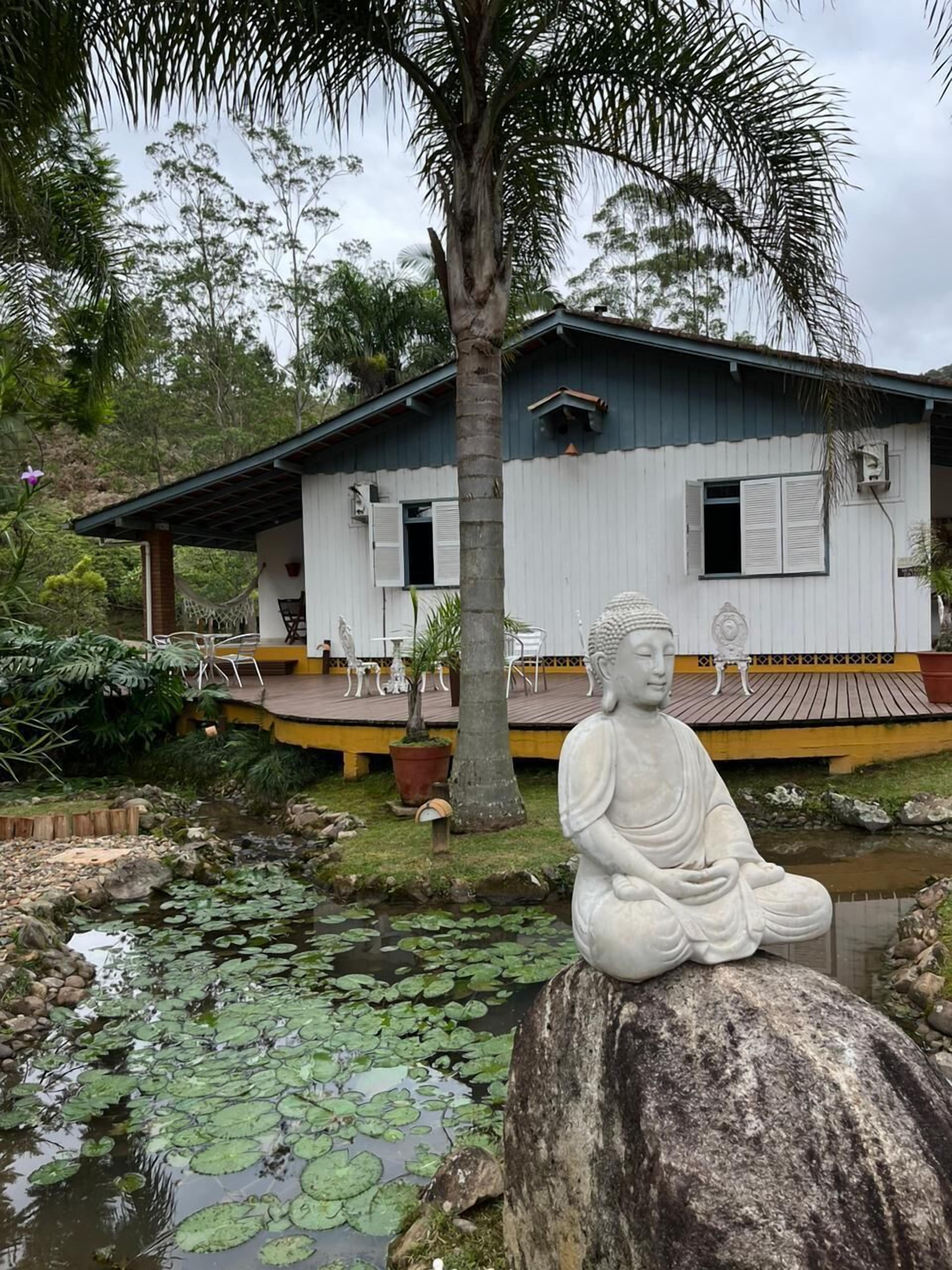Un’atmosfera tranquilla all’aperto, con una statua di Buddha su una roccia, uno stagno con ninfee e una casa con veranda e mobili da esterno.