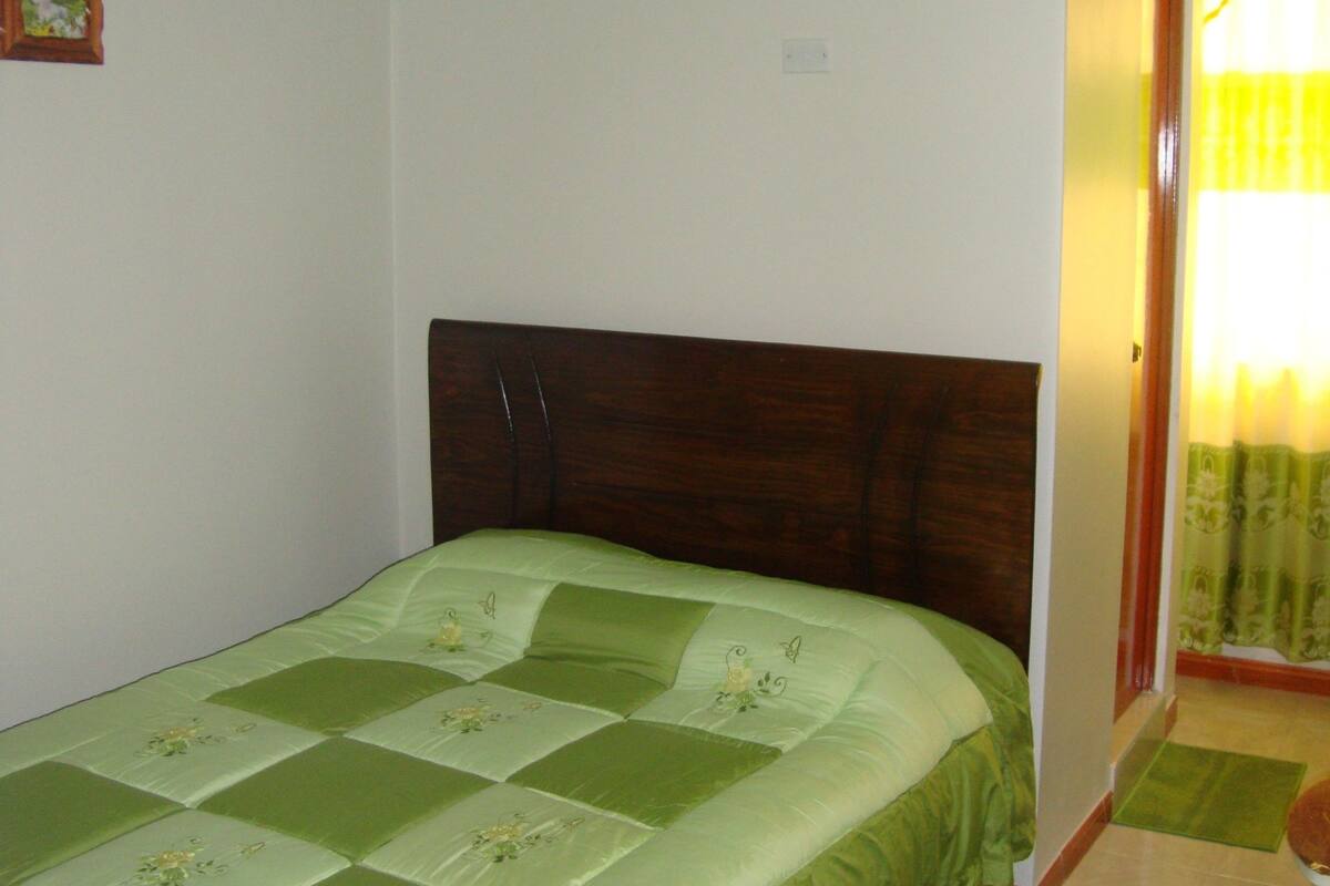A single-bed room with a green quilt, a wooden headboard, a small green rug, and a door with a yellow curtain.