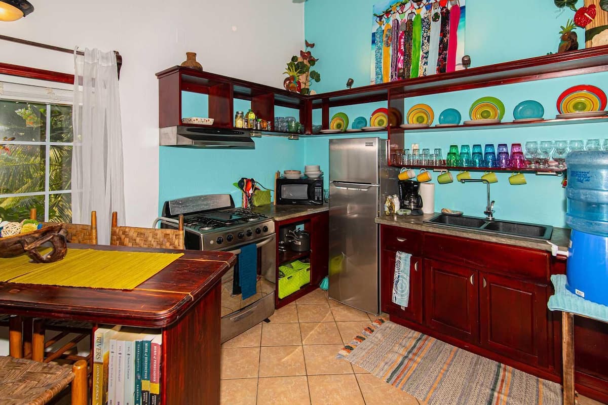 A compact kitchen with a gas stove, oven, microwave, and refrigerator. There is a dining area with a table and chairs, and a bookshelf with books.