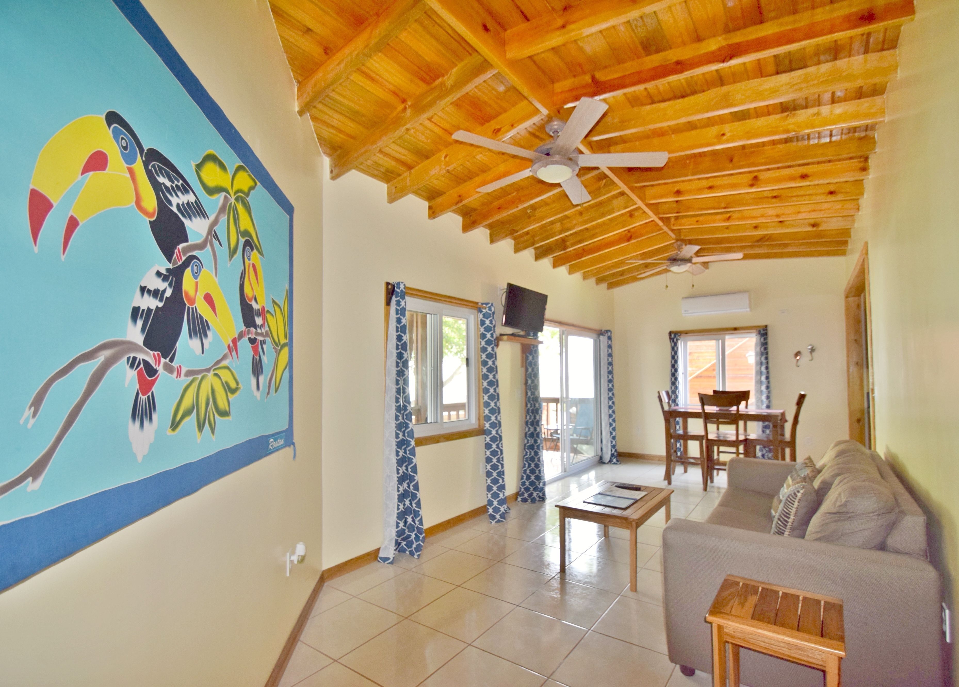 A spacious living area with a wooden ceiling, a large mural of toucans, a dining area, and a sofa.