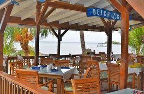A beachside restaurant with wooden tables and chairs, a covered patio, and a view of the sea. - Blue Bahia Resort (Roatan)