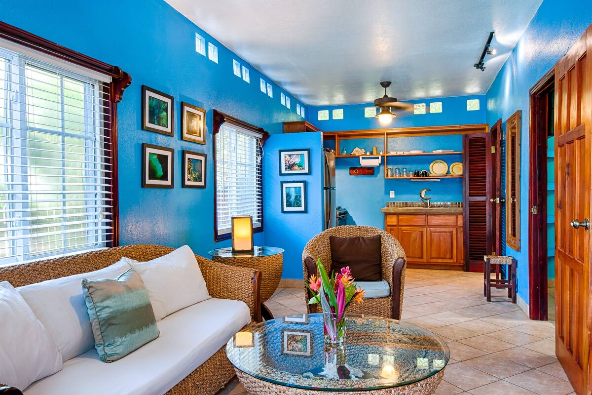 A living room with a blue wall, a white sofa, wicker chairs, a glass coffee table, and a kitchen area with wooden cabinets and a countertop.