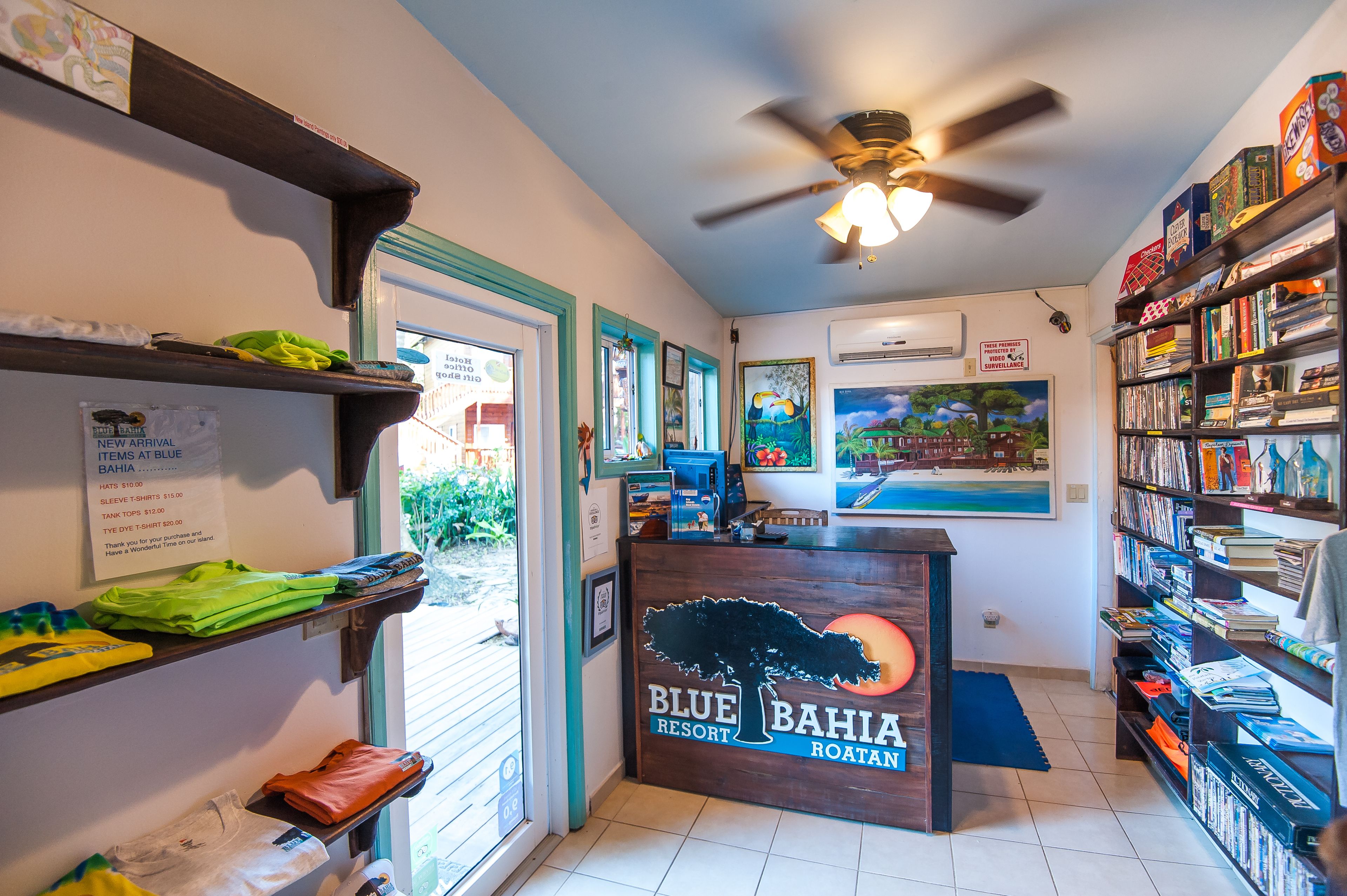 A Blue Bahia Resort Roatan reception area with a counter, shelves with folded clothes, a bookshelf, and a ceiling fan.