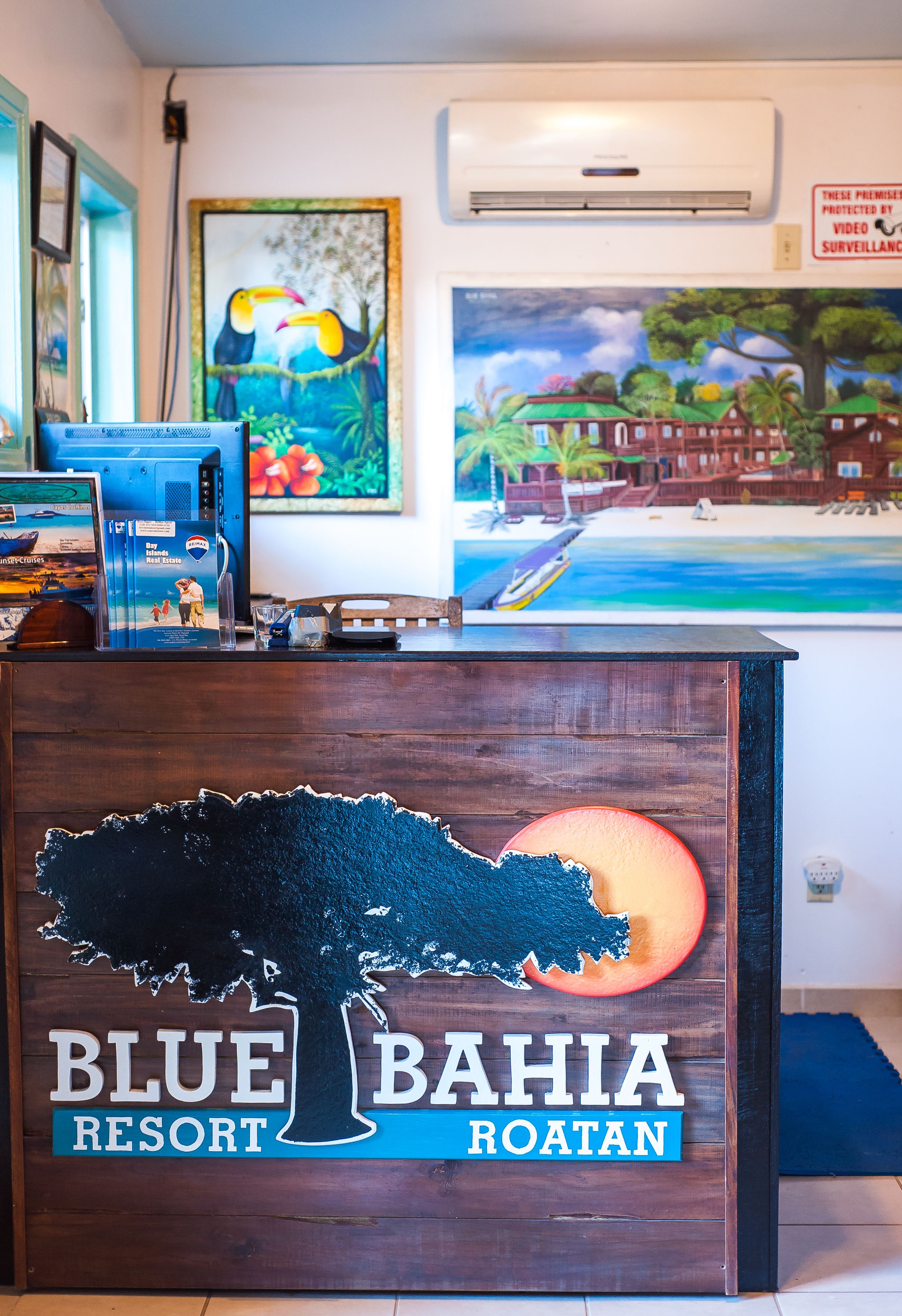 Un mostrador de recepción con un cartel que dice "Blue Bahia Resort Roatan" y una pintura de una escena tropical sobre él.