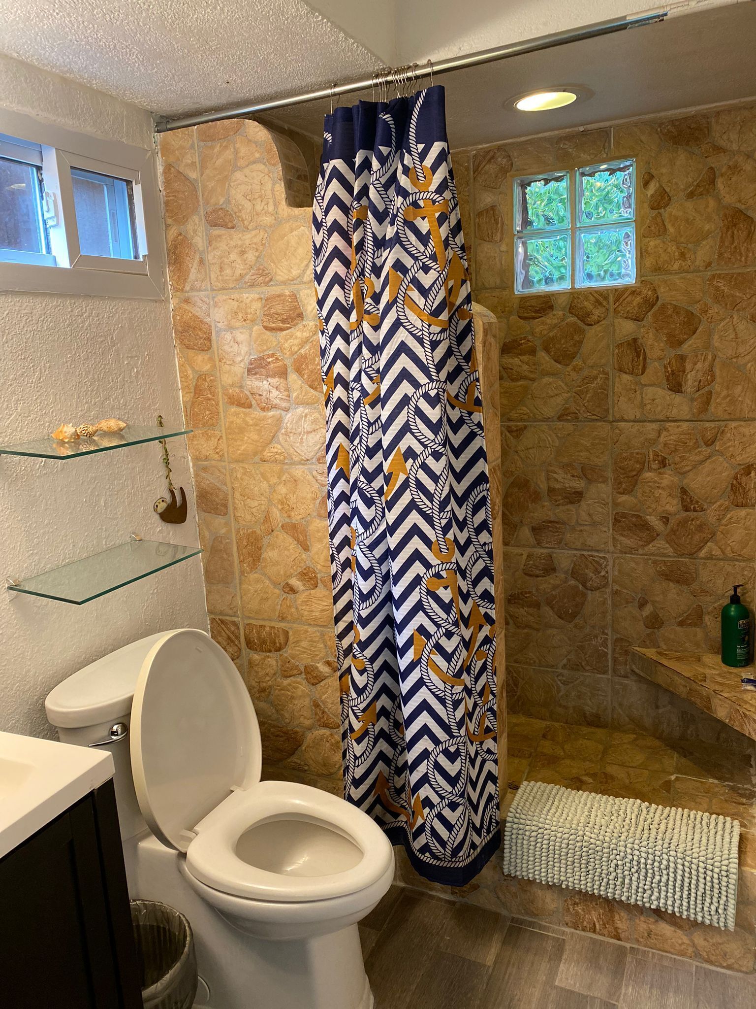 A bathroom with a stone wall, a shower curtain, a glass shelf, a toilet, and a small window.