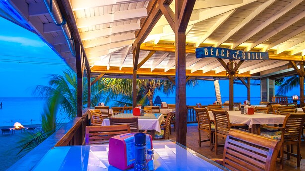 Um restaurante à beira-mar com mesas e cadeiras, um letreiro com a inscrição "BEACH & GRILL" e vista para o oceano.